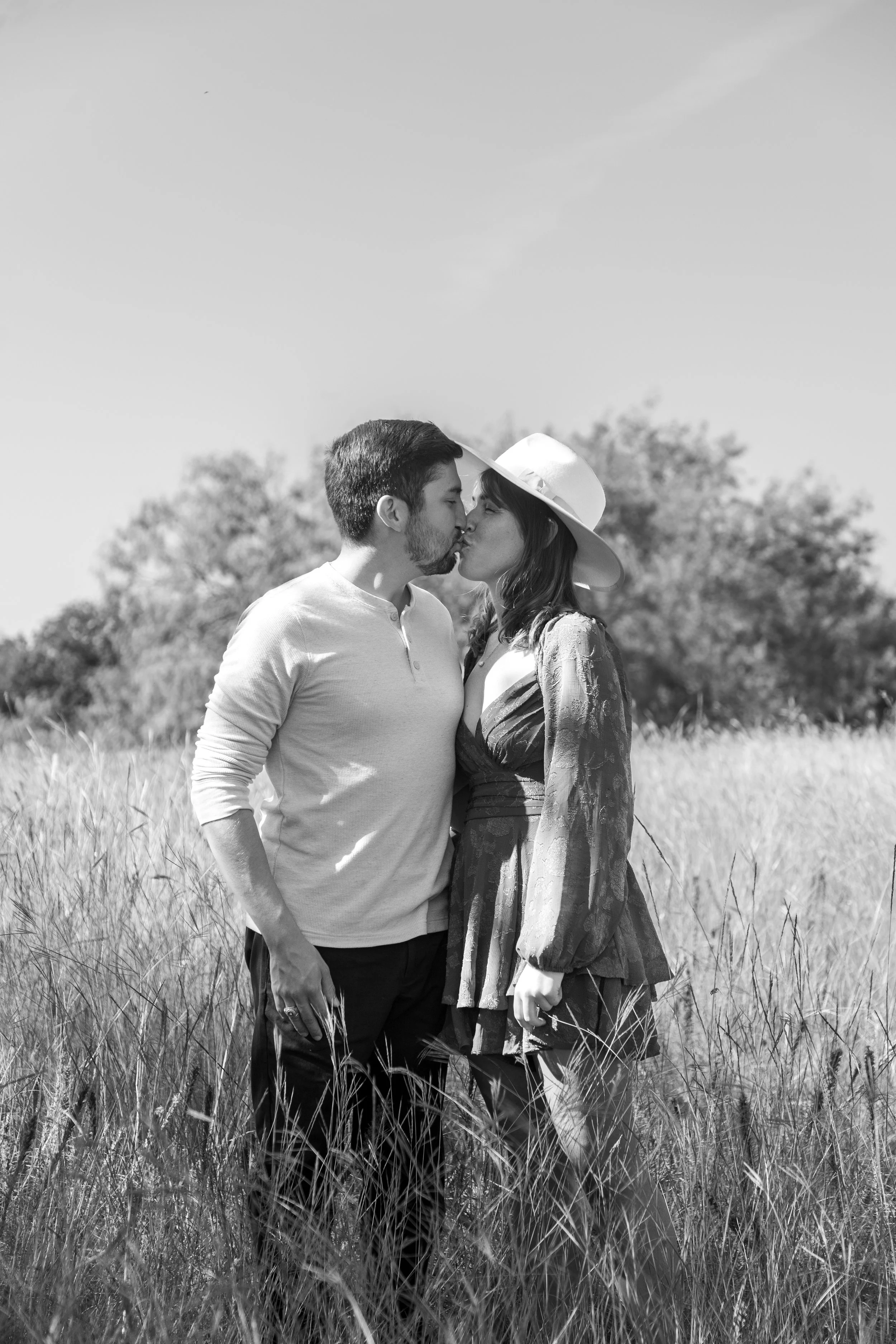 A black and white photo of a couple kissing in a grassy field, with trees in the background, the woman wearing a wide-brimmed hat.