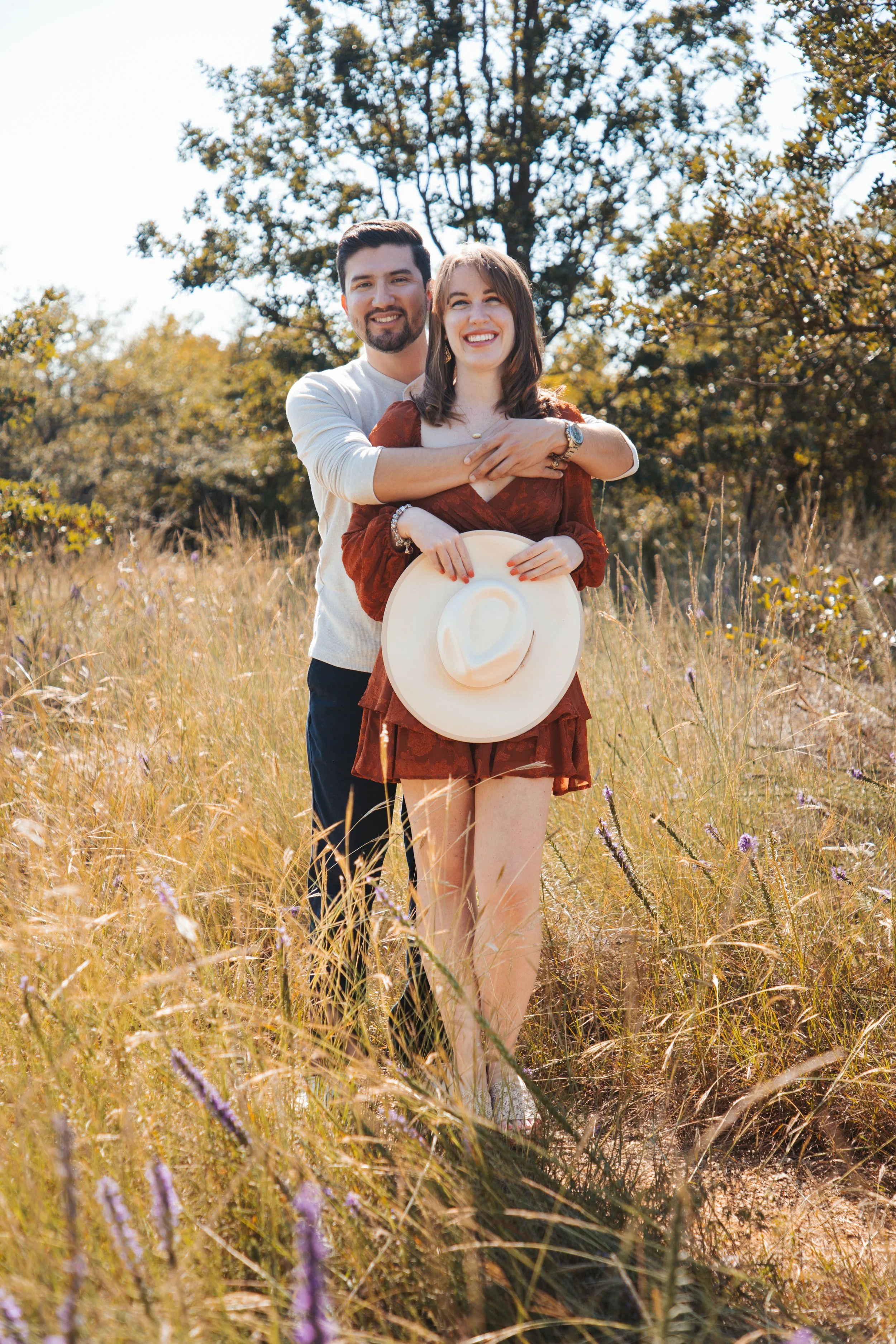 A smiling couple standing in a grassy field with trees in the background. The woman is holding a wide-brimmed hat and wearing a rust-colored dress. The man is hugging her from behind, wearing a light-colored shirt.