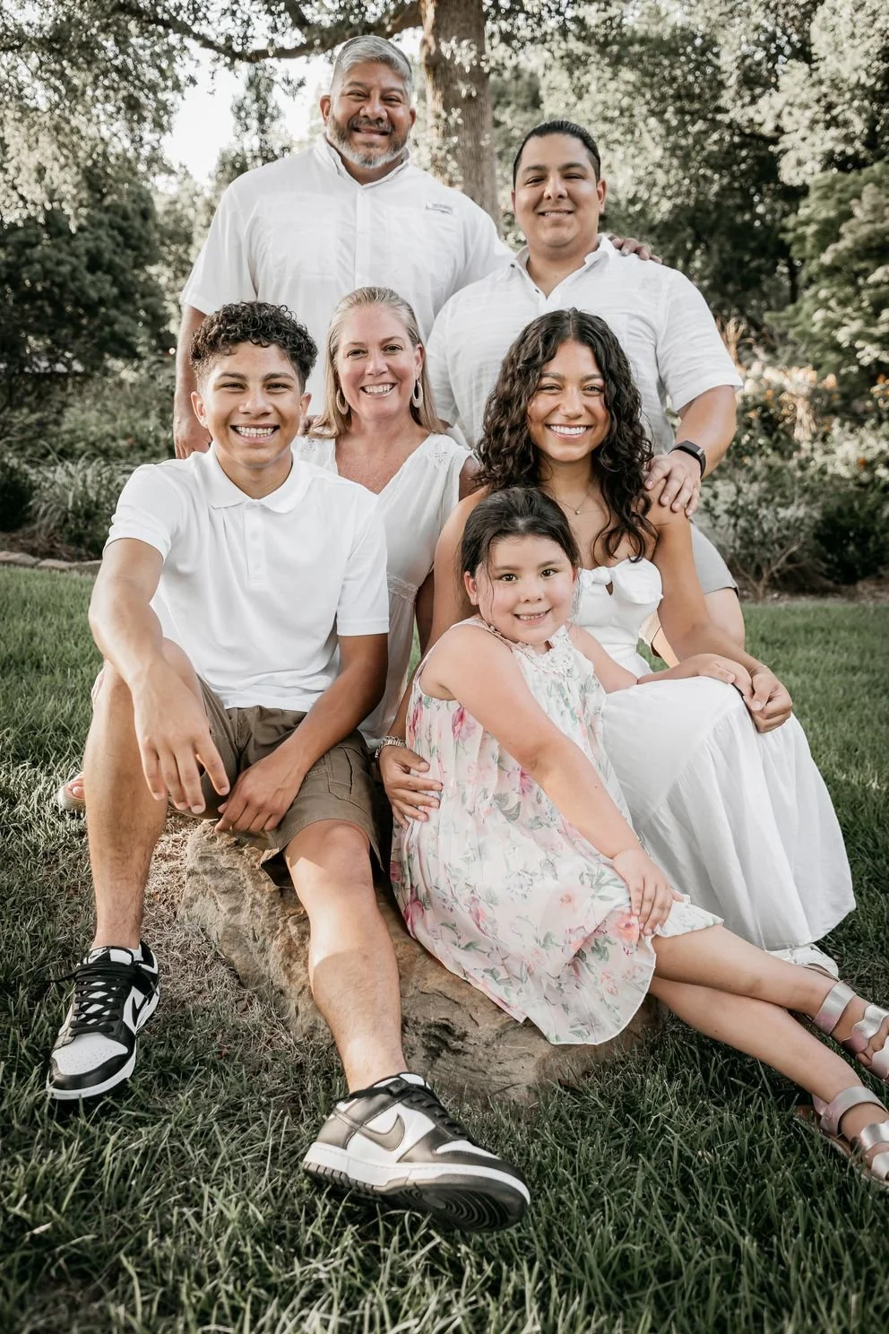 Family photo outdoors with six people, seated and standing on grass and a large log, surrounded by trees and greenery, smiling at the camera.