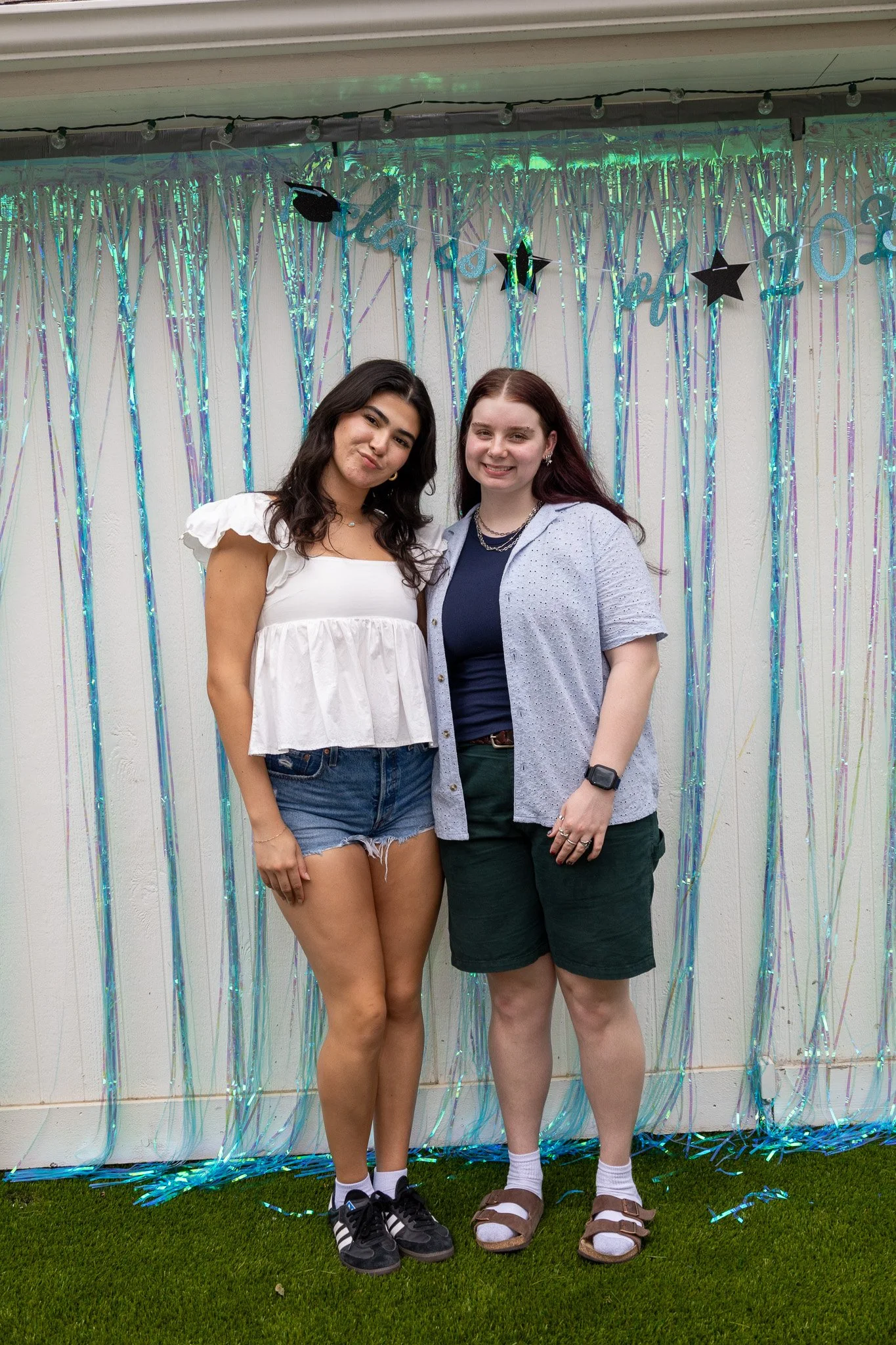 Two women standing together in front of a shiny, colorful tinsel curtain backdrop at a celebration, smiling at the camera.