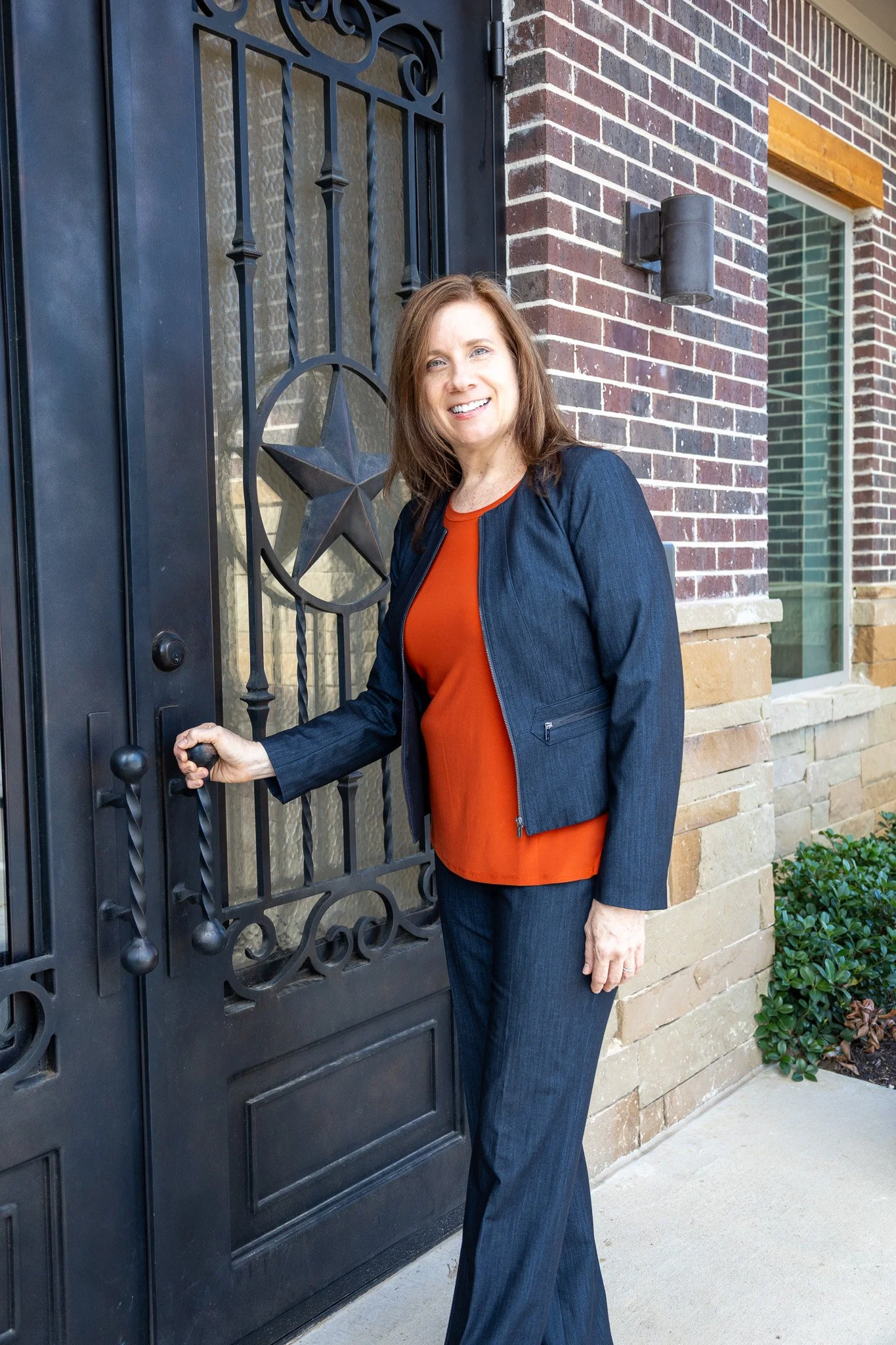 A woman with brown hair, wearing a dark blue jacket and pants, and an orange top, standing outside a house with a black metal gate featuring a star design. She is holding the gate handle and smiling.