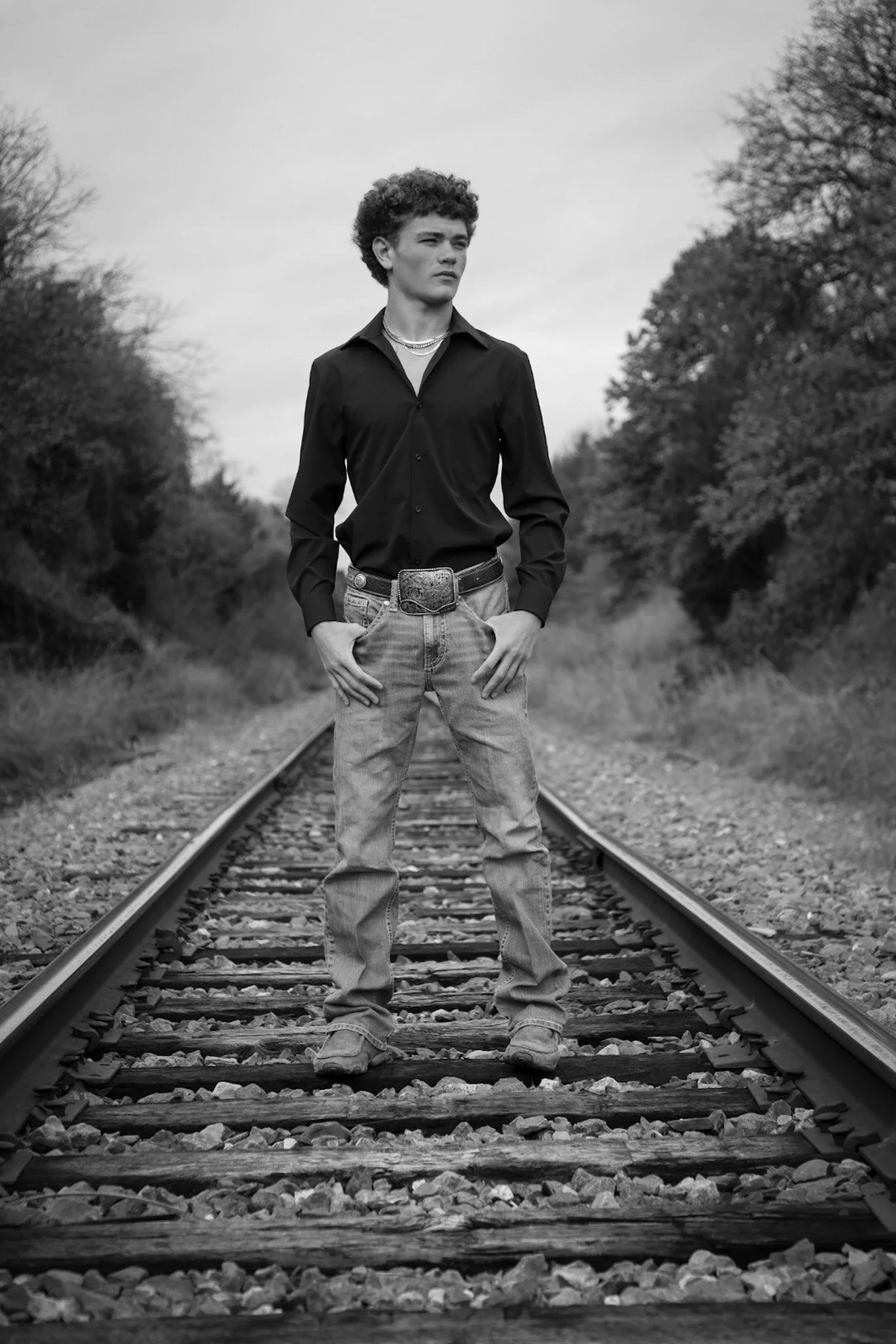 A young man with curly hair stands on railroad tracks surrounded by trees, wearing a black button-up shirt, jeans, and jewelry, in black and white photography.