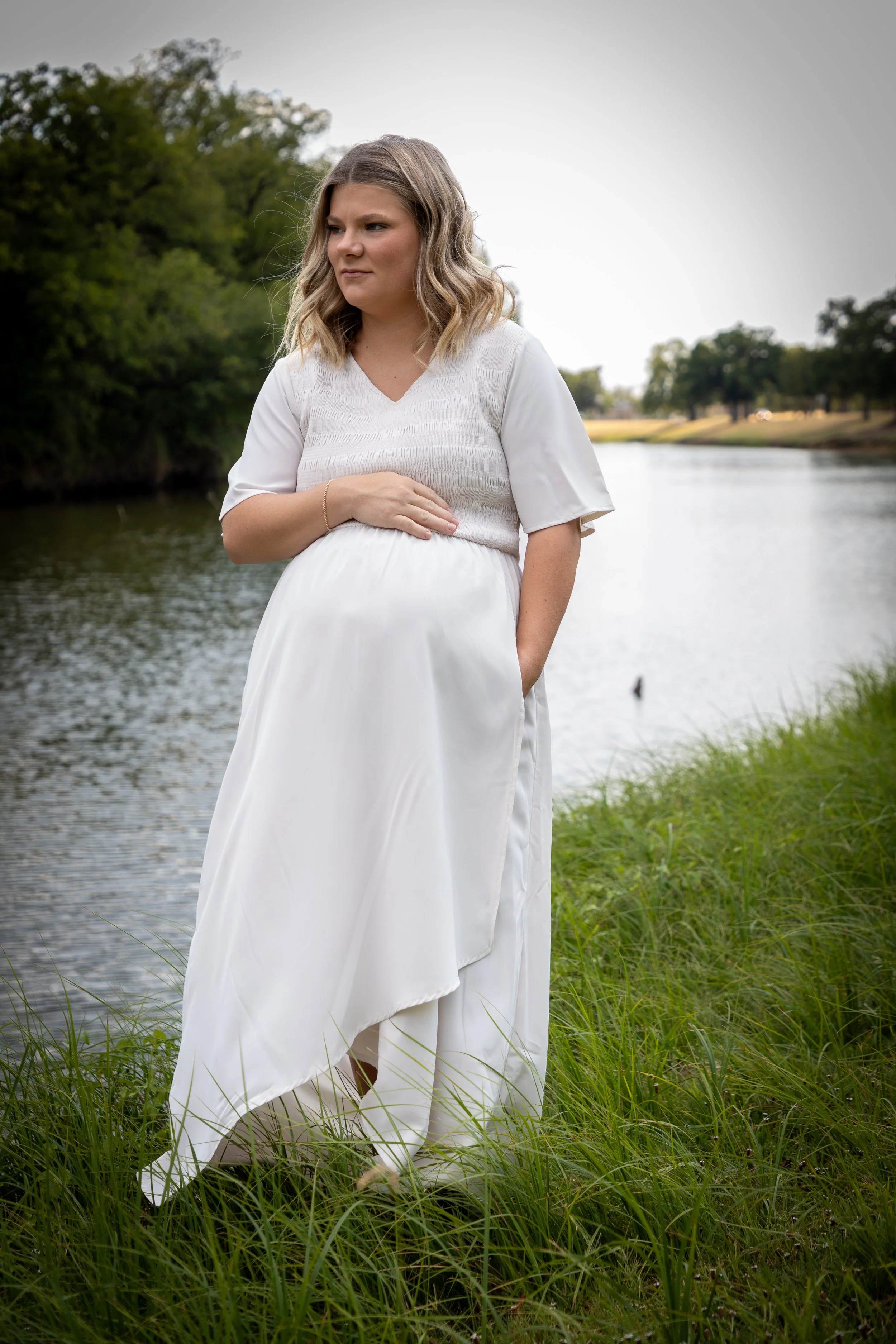 A pregnant woman in a white dress standing outdoors by a river, with green grass and trees in the background.