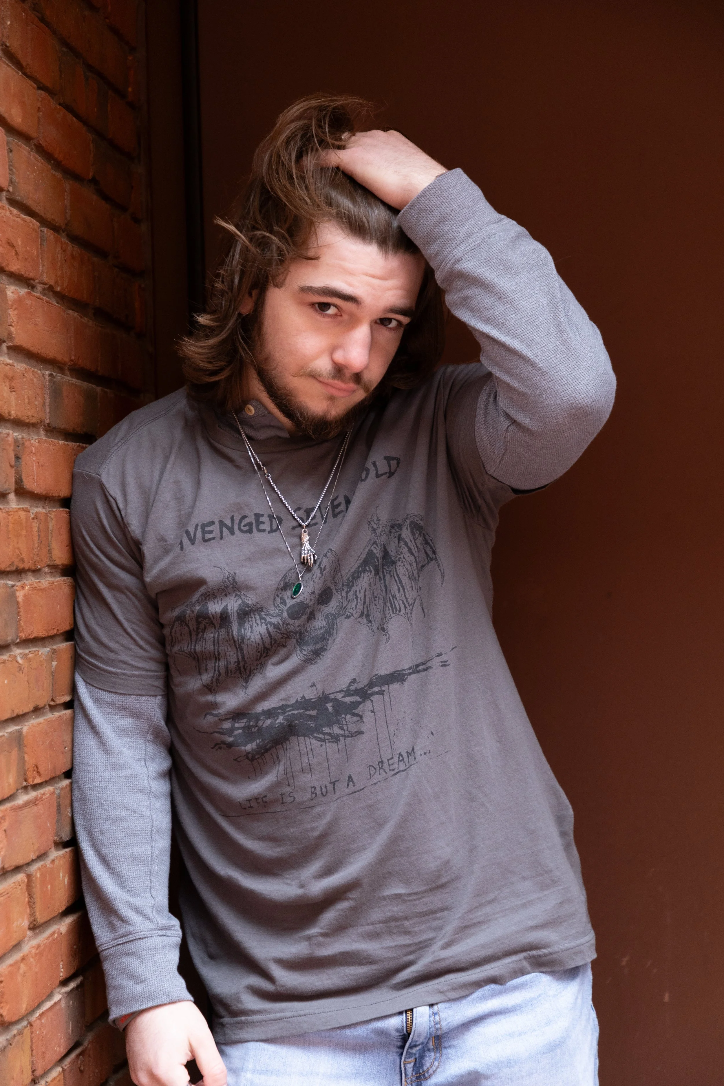 A young man with long hair, beard, and mustache posing with his hand in his hair against a brick wall and brown background.