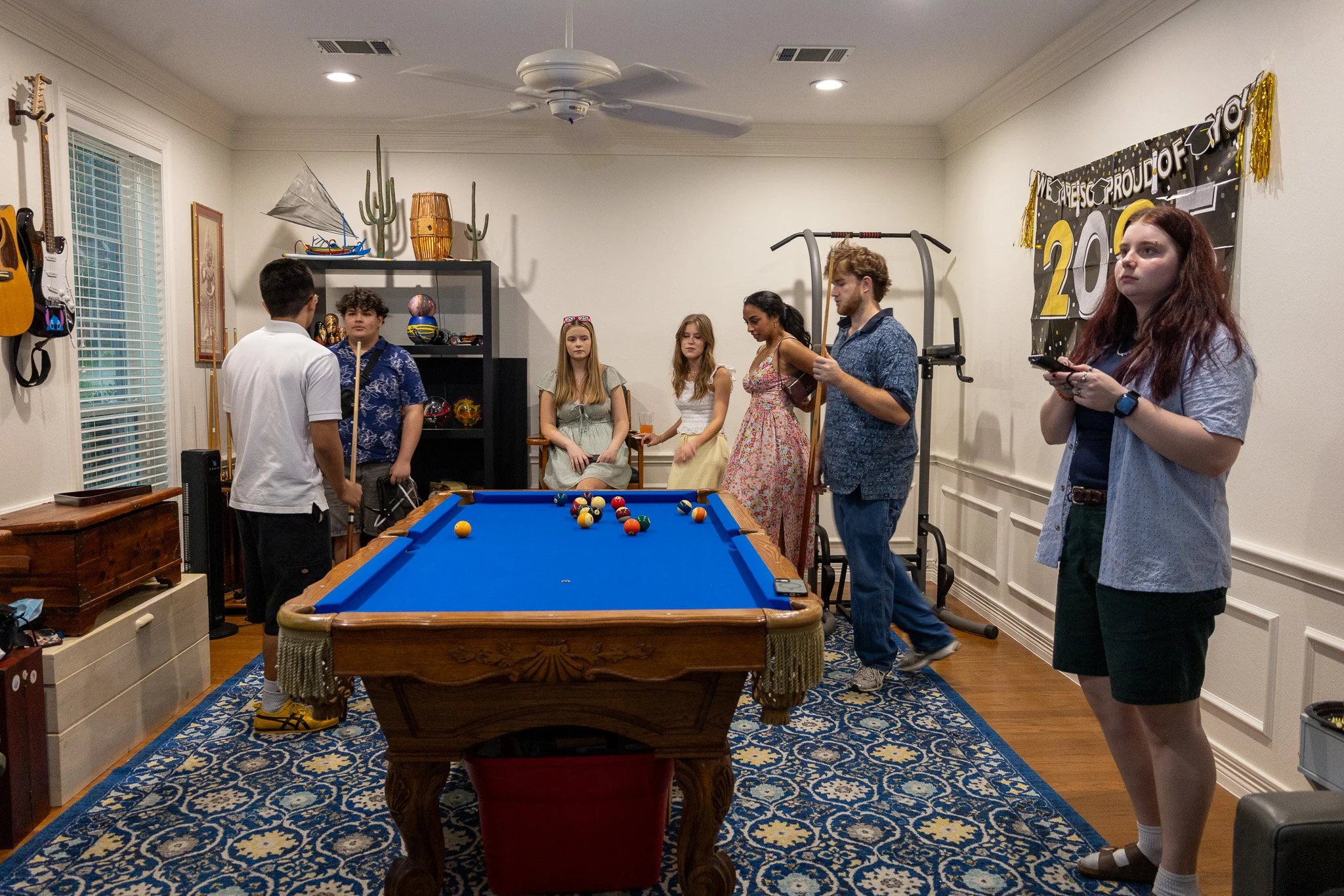 A group of young adults and teenagers gathered around a blue pool table in a decorated living room, some talking and one girl on her phone, with musical instruments and sports equipment visible in the background.
