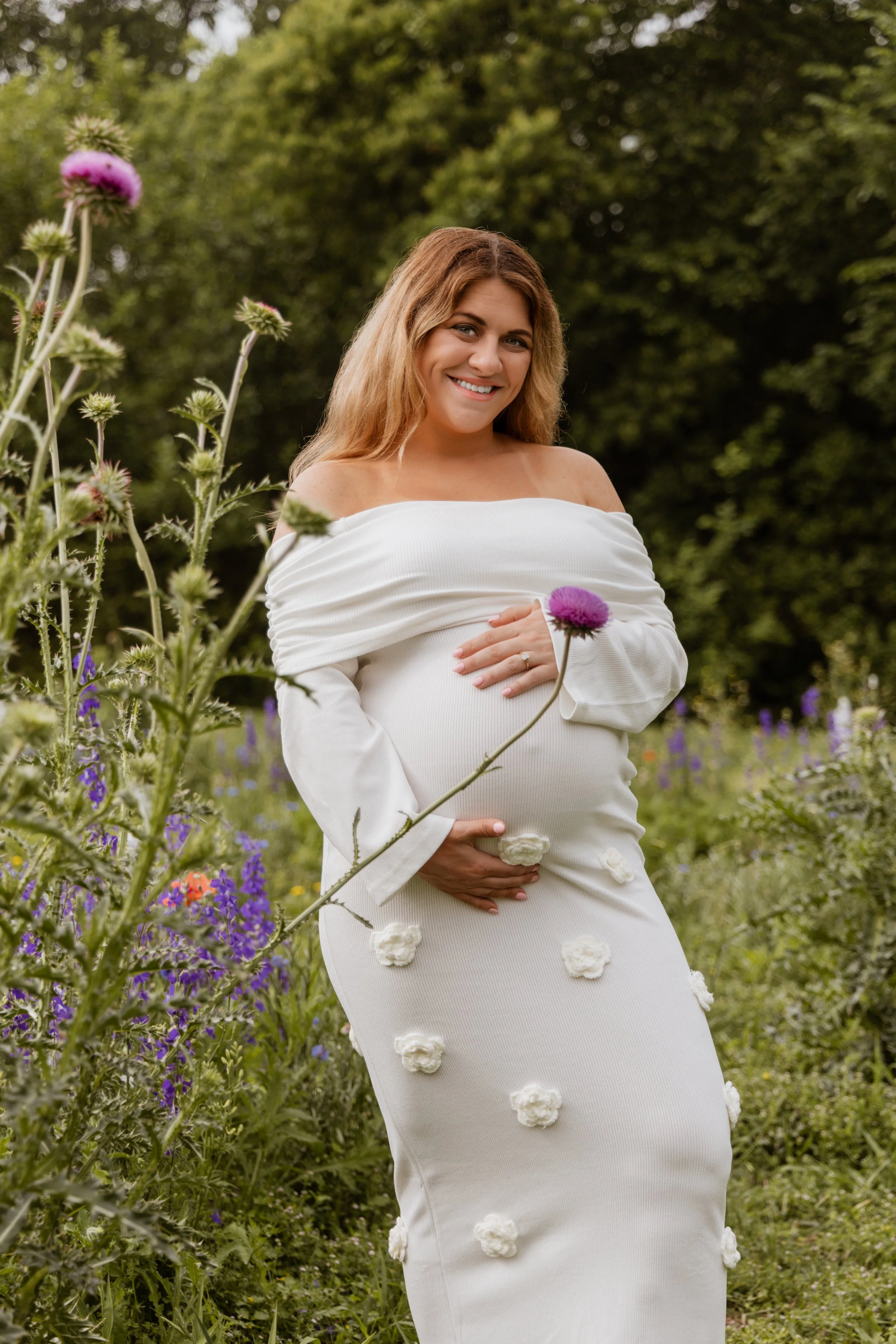 A pregnant woman smiling and holding her belly in a garden with purple and green flowers and trees.