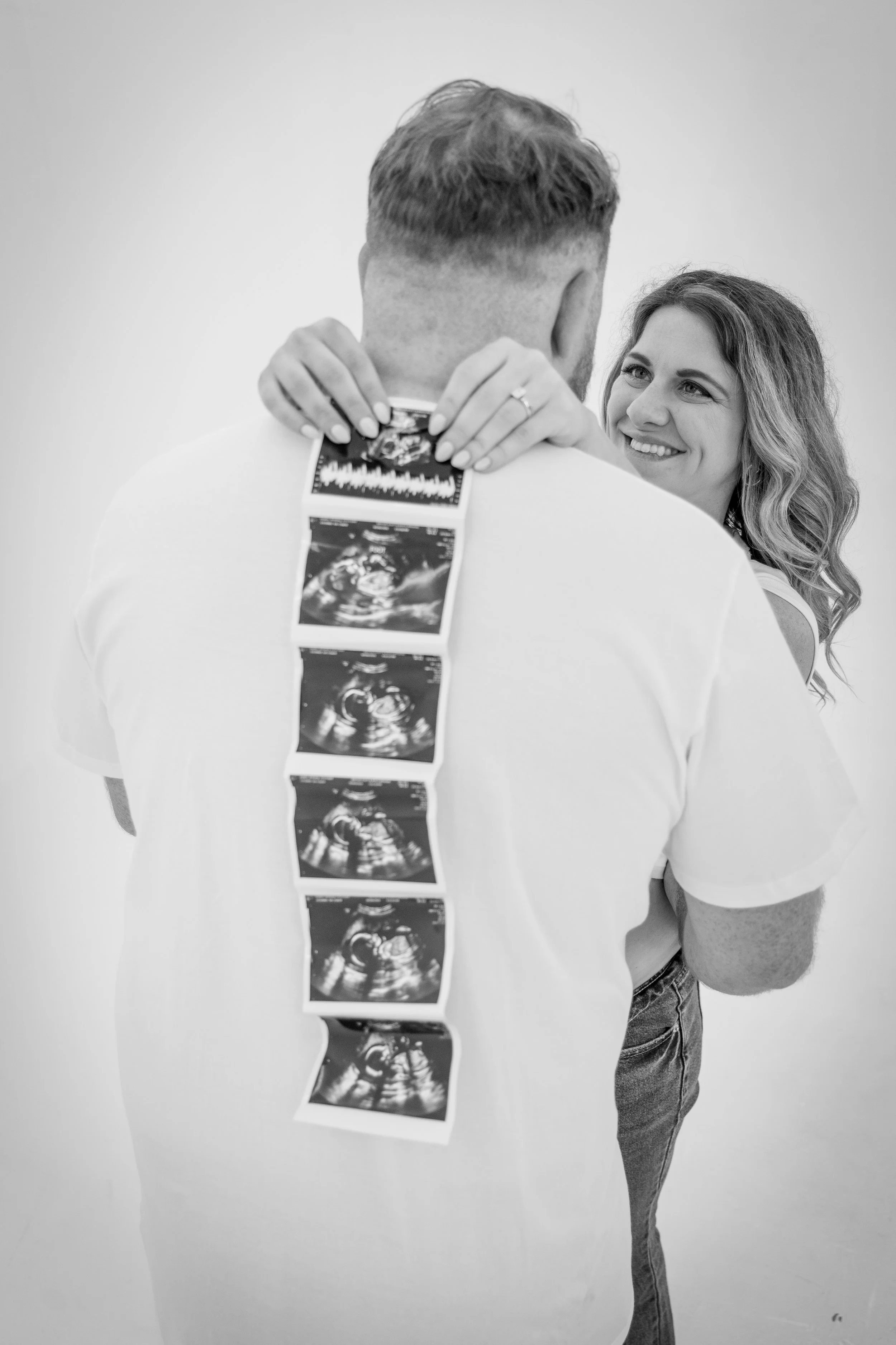 A woman with long wavy hair smiling at a man with short hair, who is holding ultrasound images on his back with his hands, in front of a plain light-colored background.