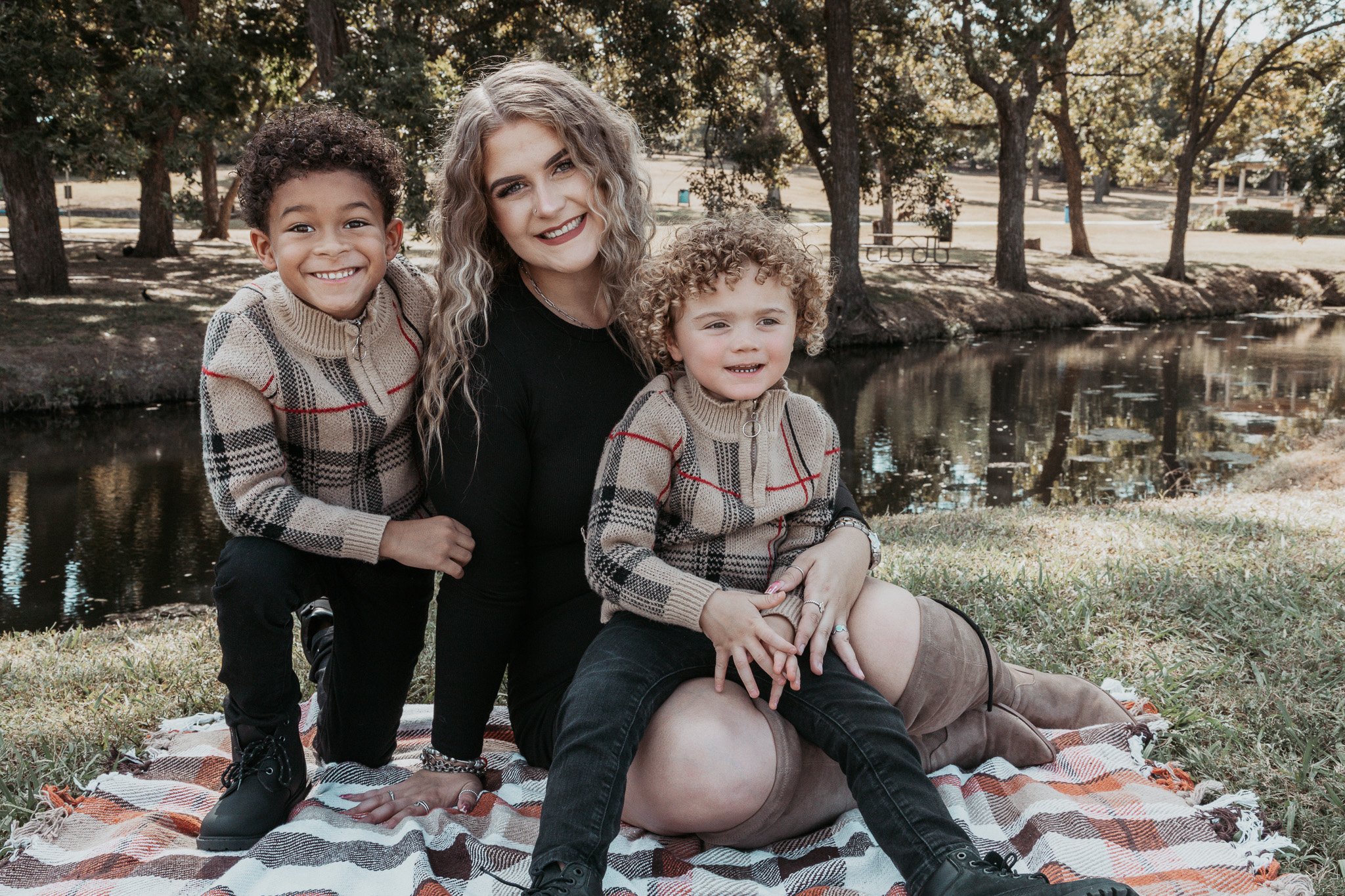 A woman and two young boys with curly hair sitting on a blanket lakeside in a park, all smiling.