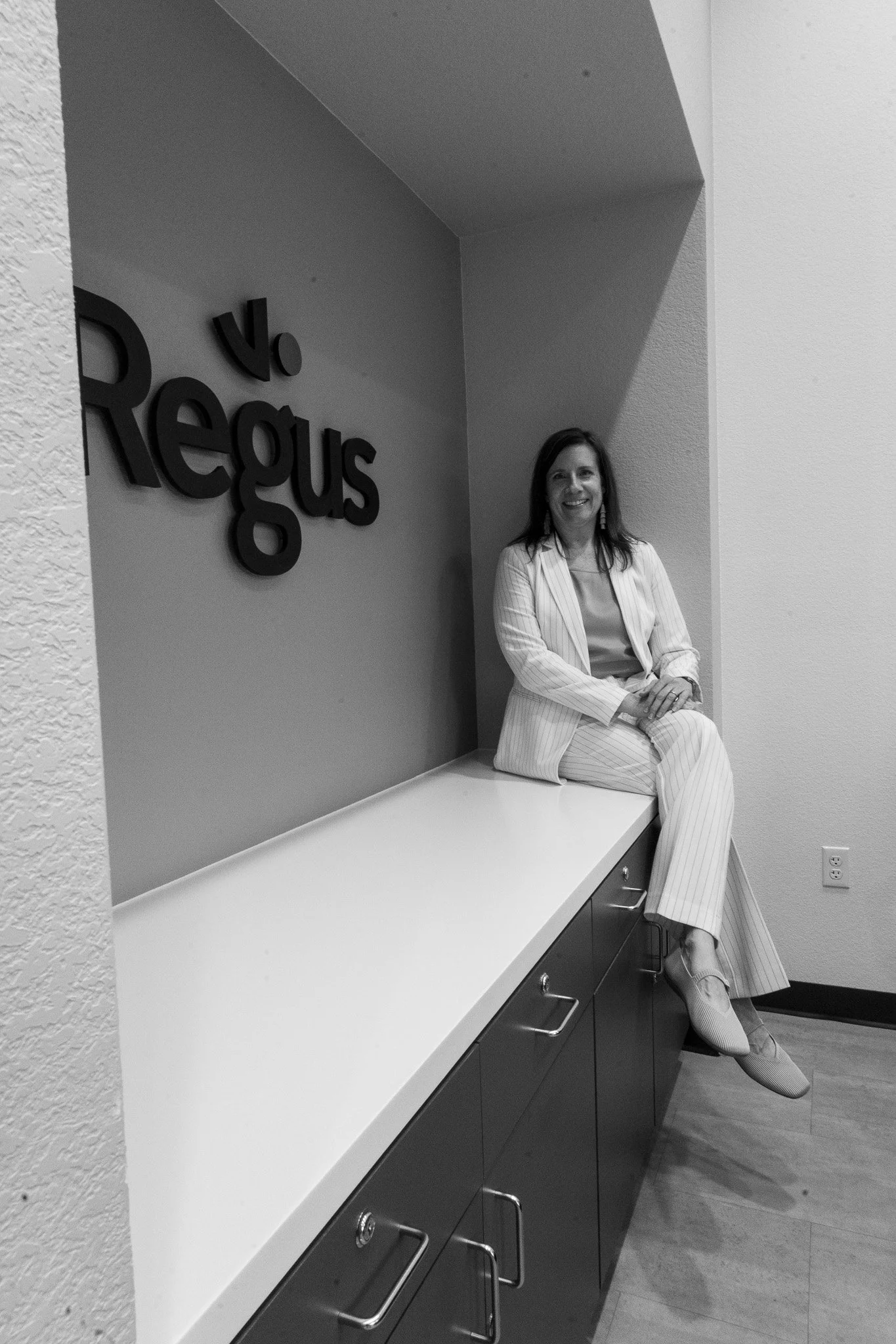 A woman in a white pinstripe suit seated on a white countertop in an office setting, smiling, with a wall sign that reads 'V Regus' behind her.