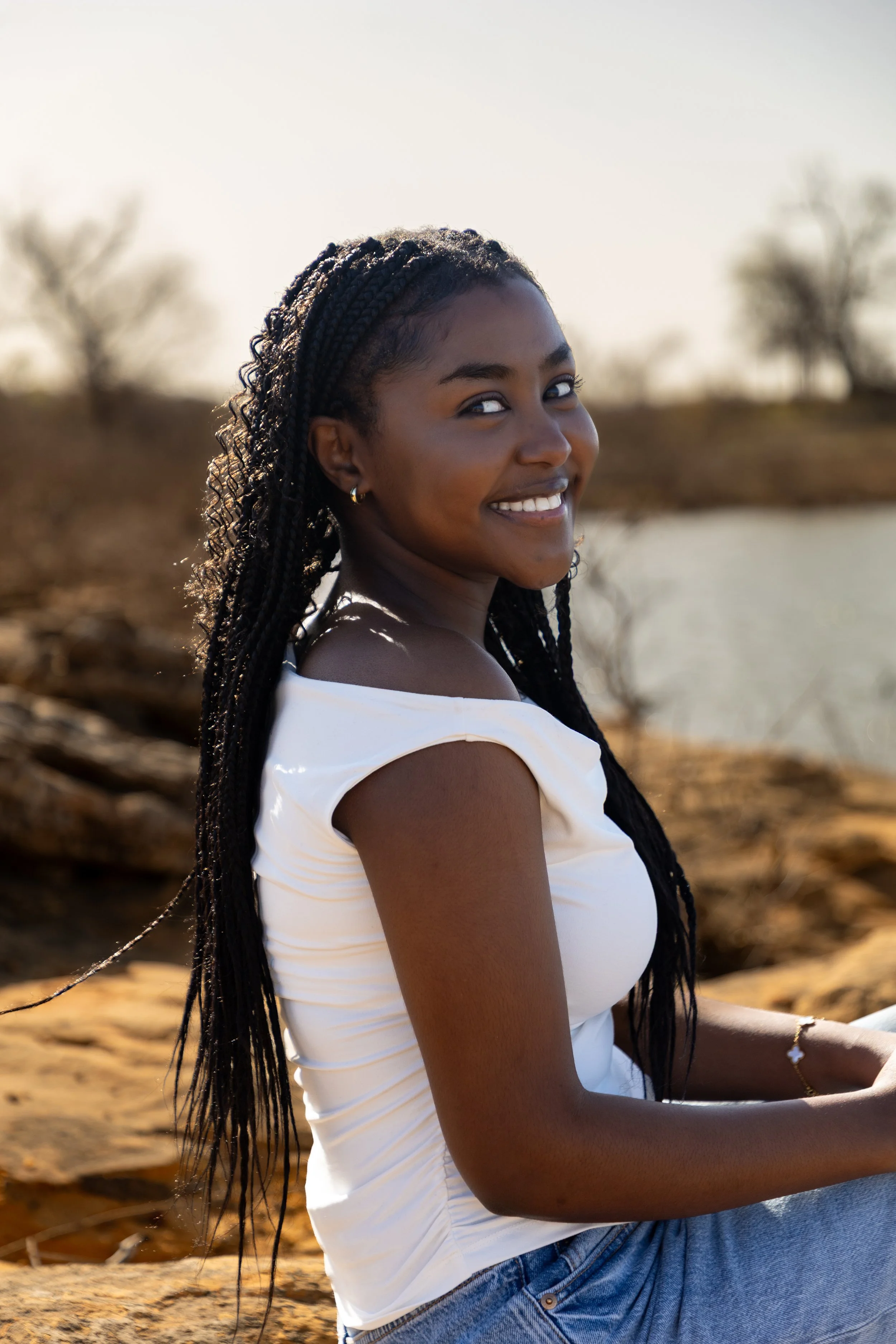 A young woman with long braided hair sitting outdoors near a body of water, smiling at the camera, wearing a white top and blue jeans in a natural setting.