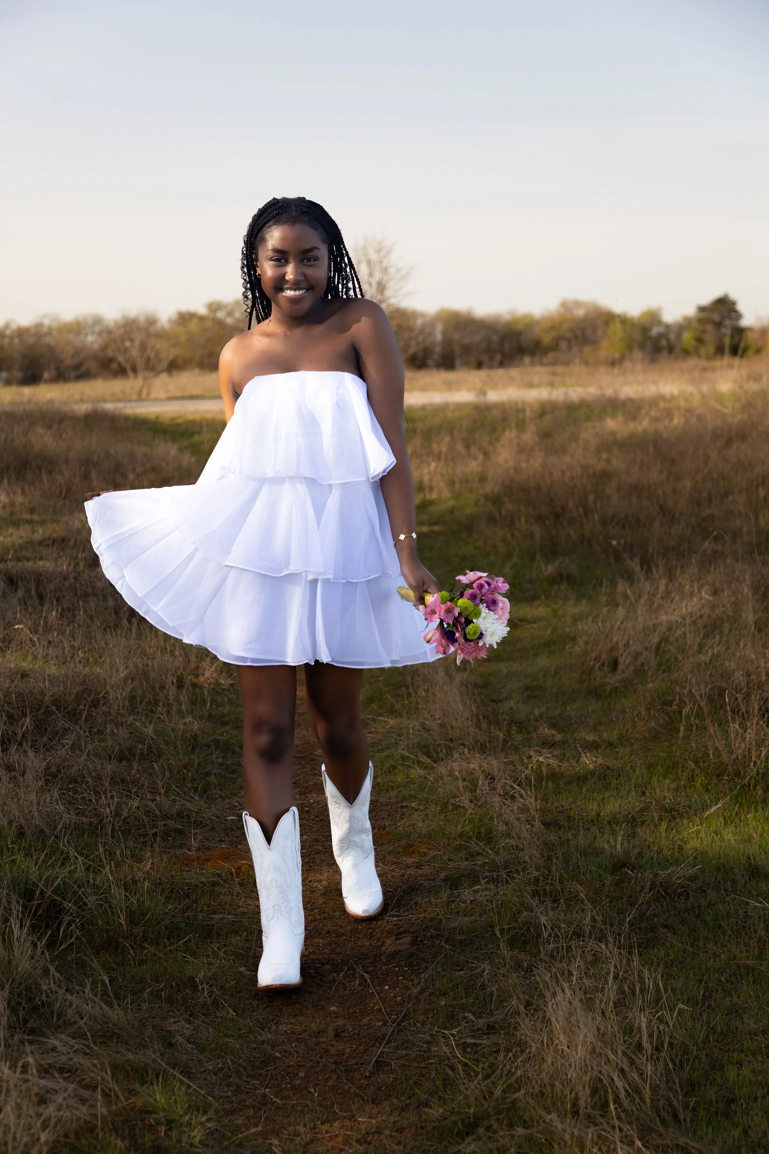 A young girl smiling in a white strapless dress and cowboy boots, holding a bouquet of pink, white, and green flowers, standing on a grassy trail outdoors during daytime.