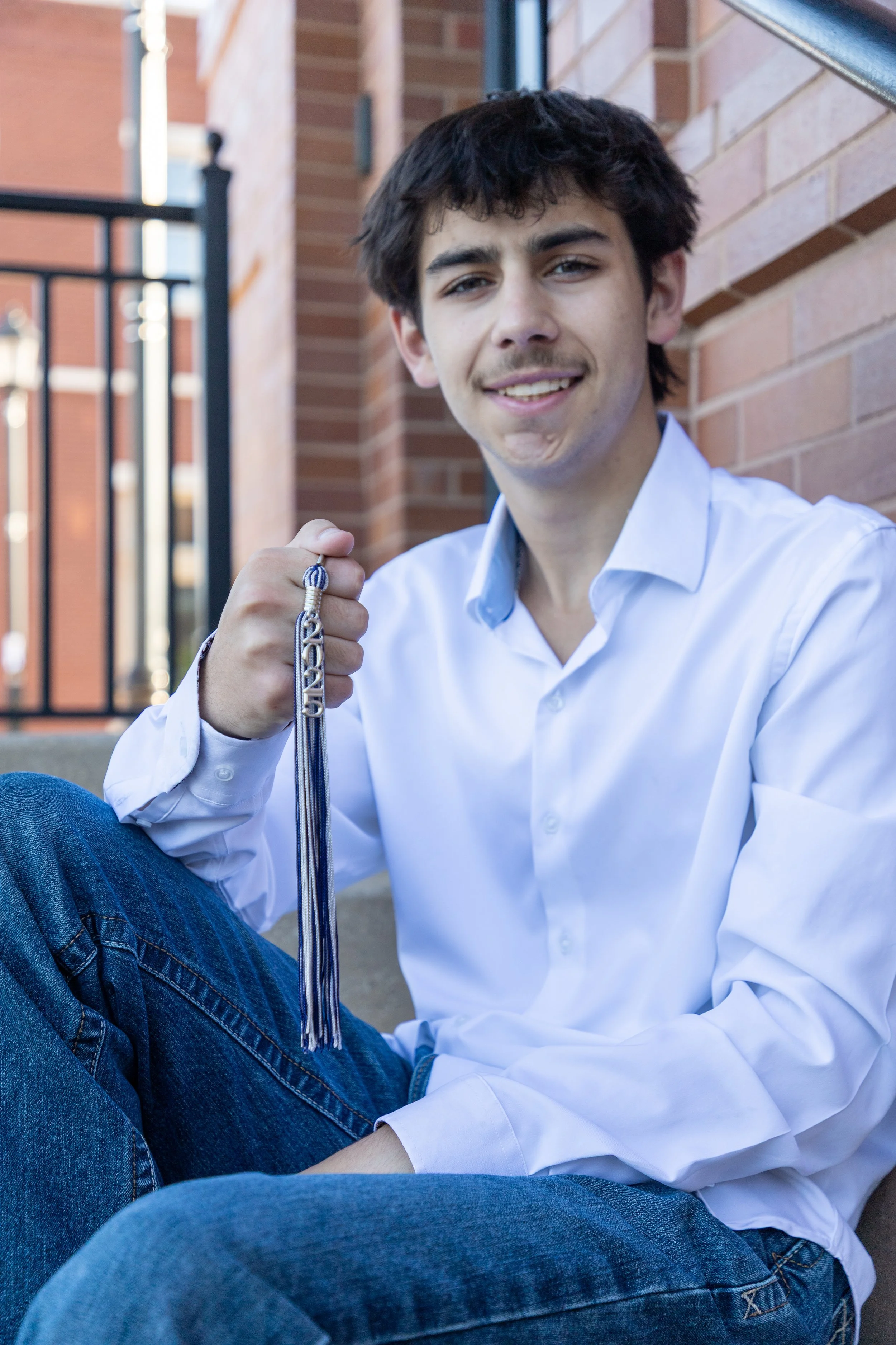 A young man with dark brown hair, wearing a white button-up shirt and blue jeans, sitting on steps outside next to a brick wall, holding a silver medal with a striped ribbon, smiling at the camera.