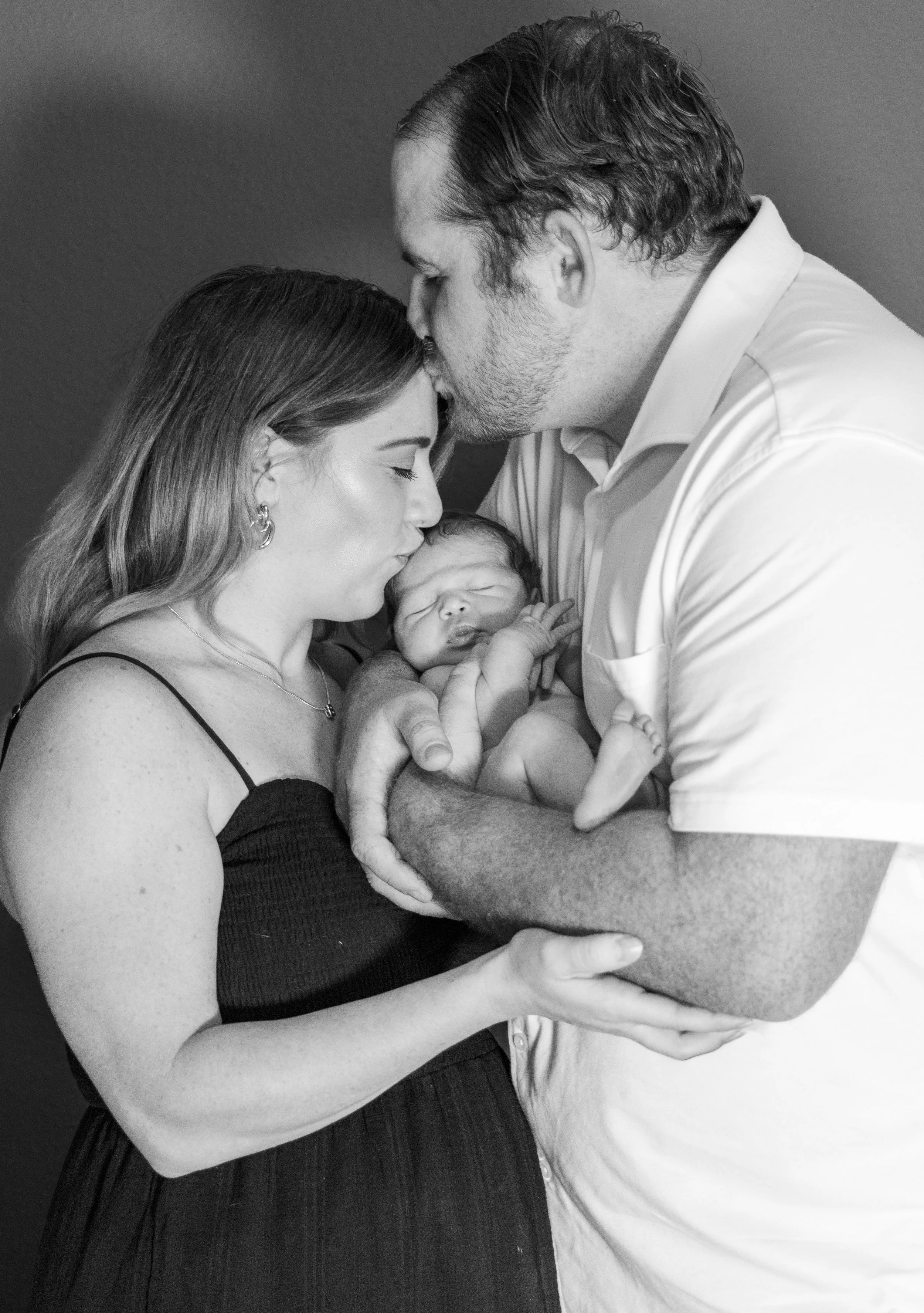 A couple holding their newborn baby close, with both adults kissing the baby's forehead, sharing a tender moment in black and white.