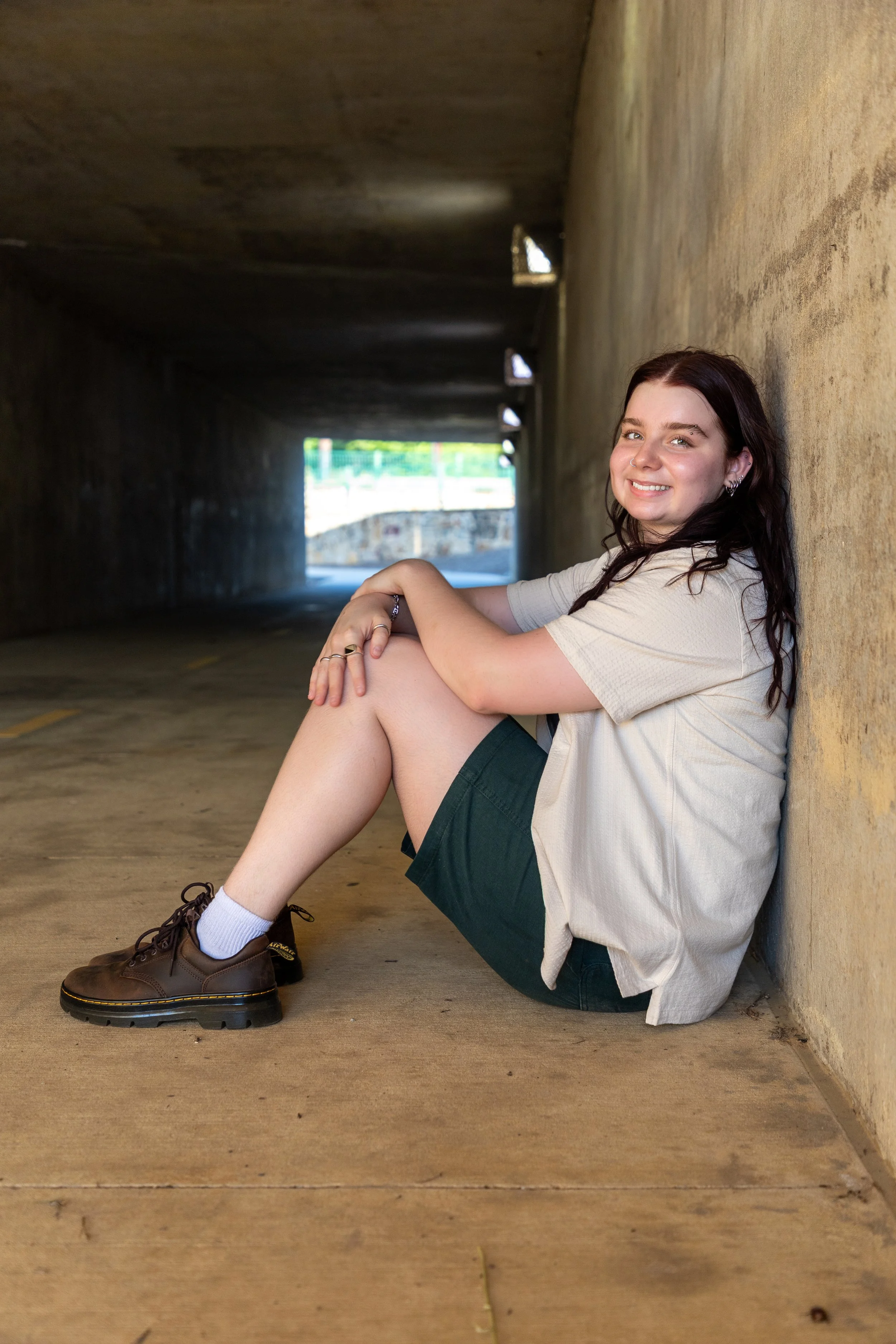 A young woman with dark hair, wearing a beige shirt, green shorts, white socks, and brown shoes, sitting on the ground and leaning against a yellow wall in a tunnel with an opening to sunlight at the end.