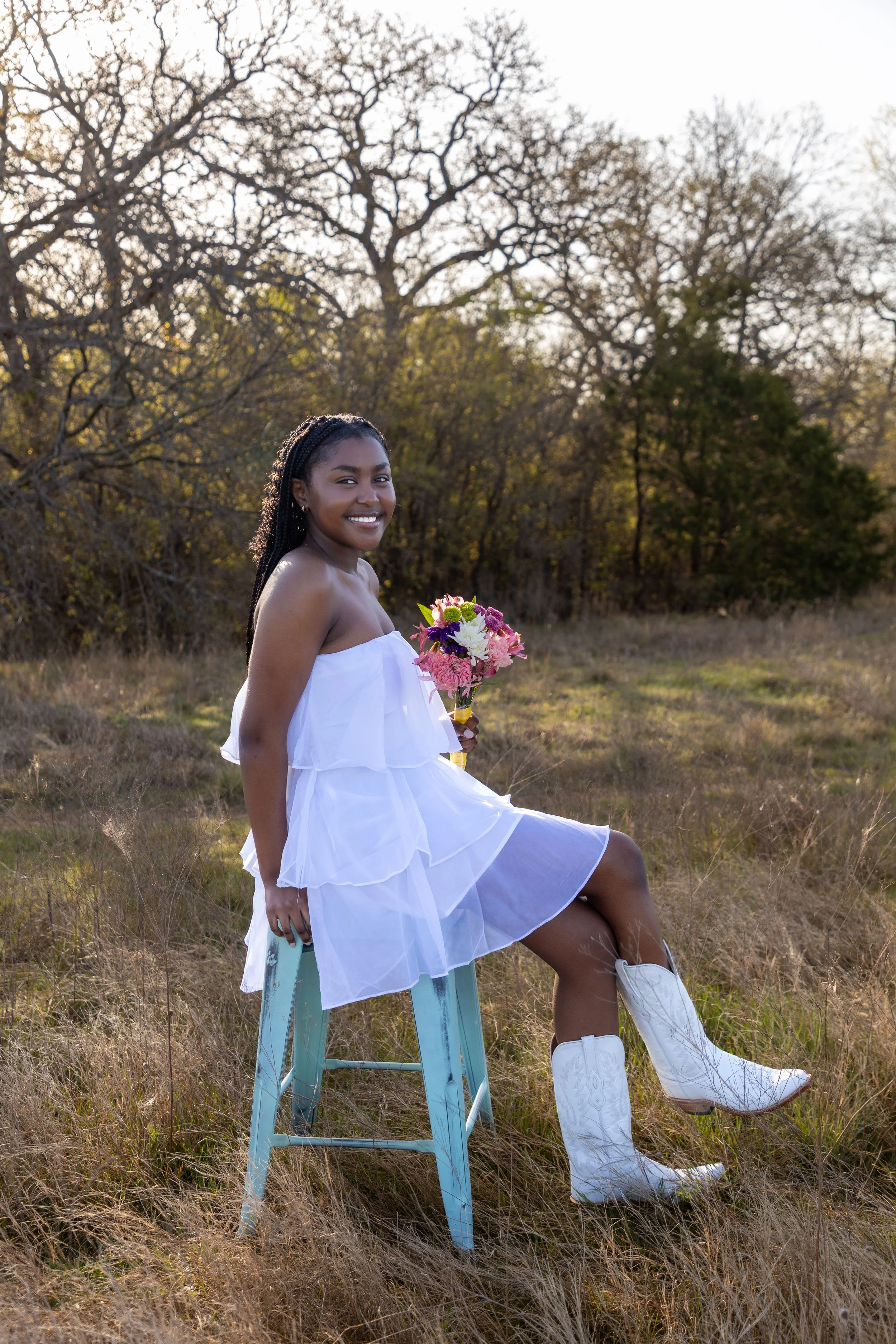 A woman in a white dress sitting on a turquoise stool in a grassy field holding a colorful bouquet of flowers, with trees and a cloudy sky in the background.