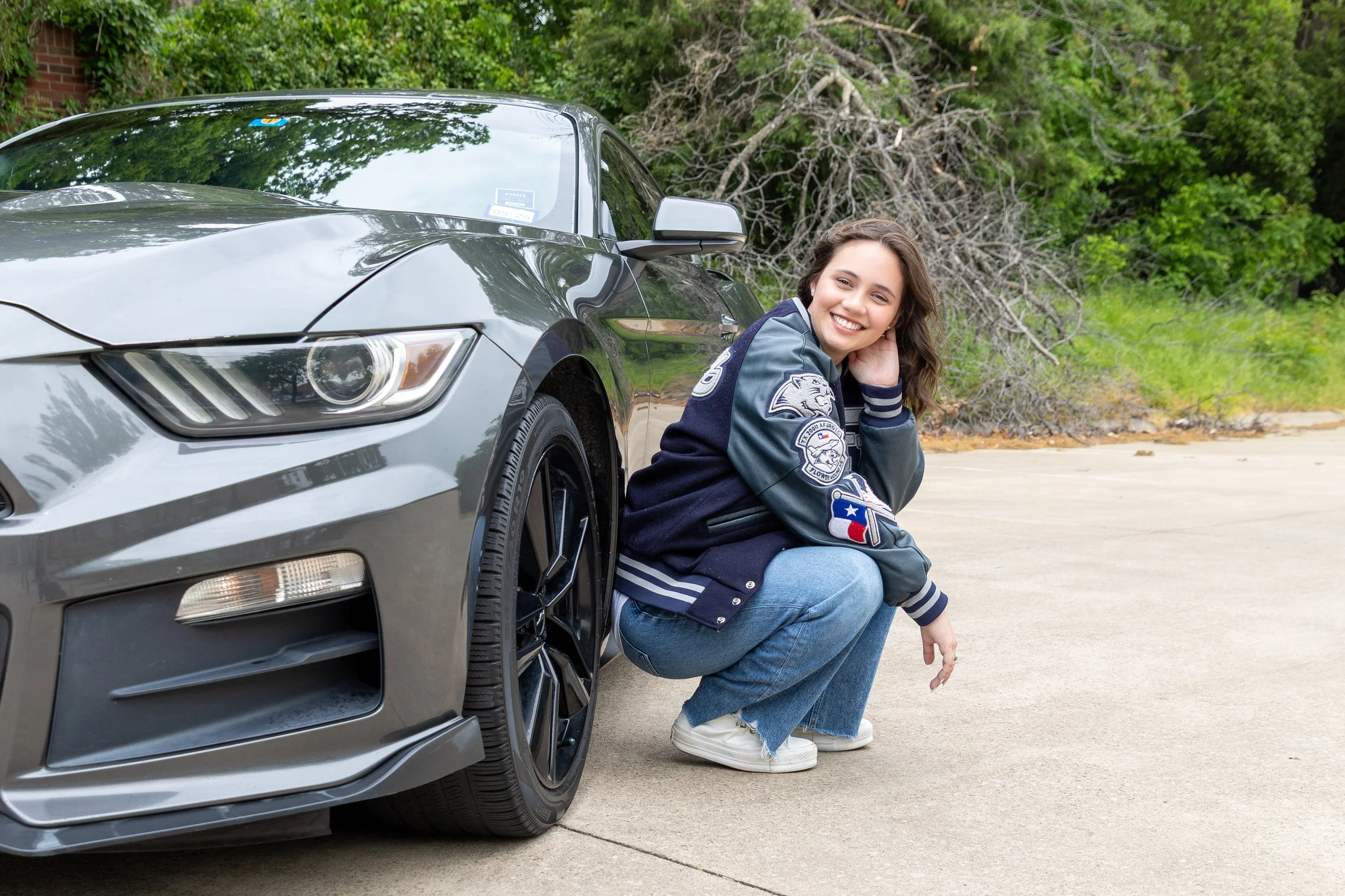 A smiling young woman in a navy varsity jacket and jeans squatting next to a gray sports car on a concrete driveway with trees and greenery in the background.