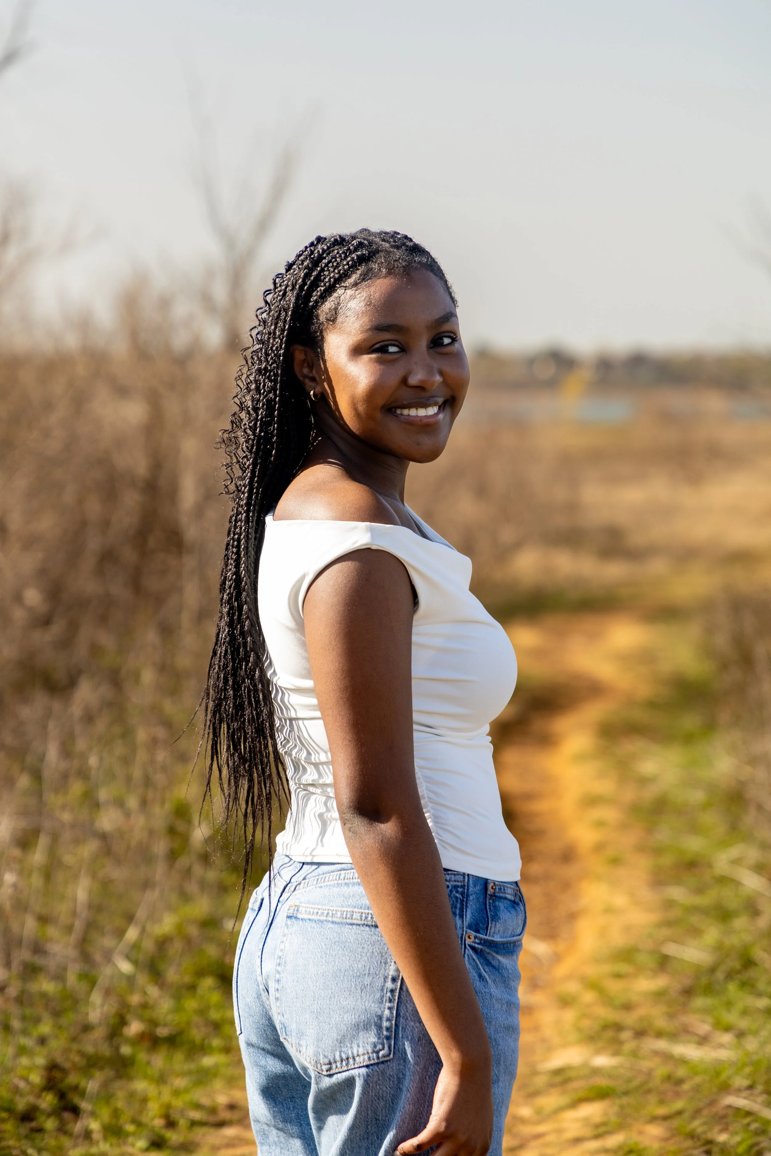 A young woman with dark braided hair smiling outdoors on a dirt path with dry bushes in the background.
