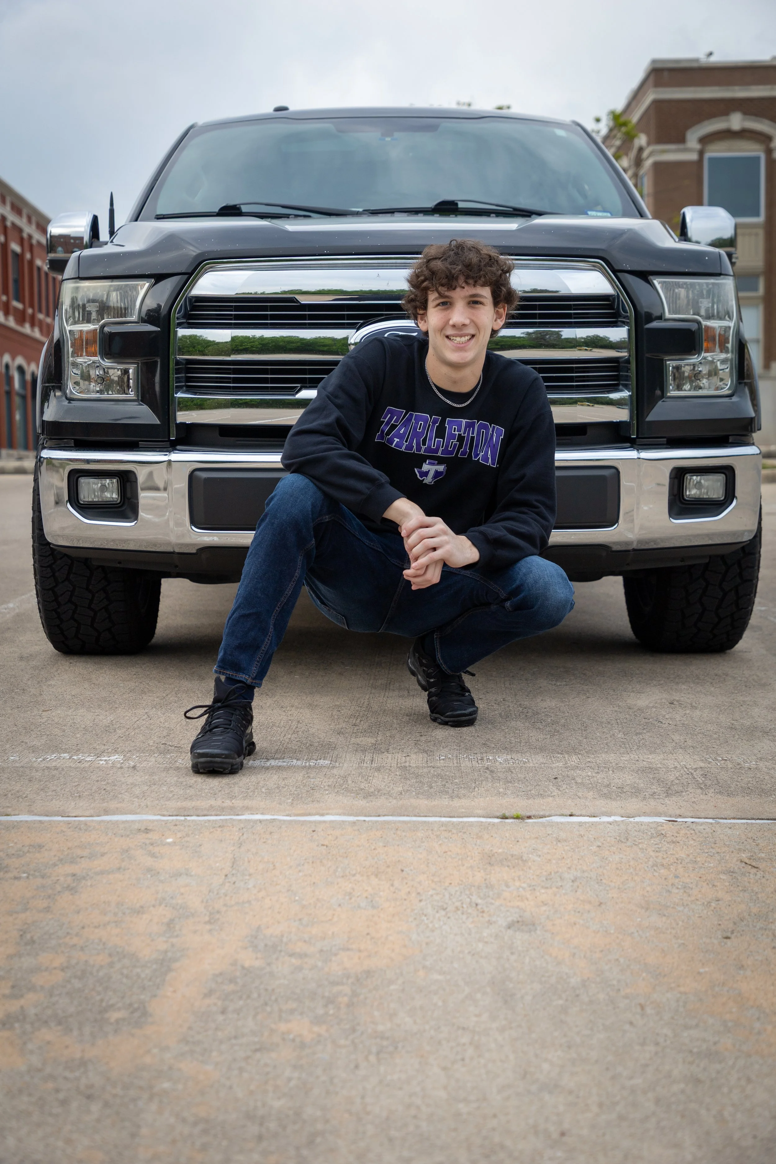 Young man with curly hair crouching in front of a black pickup truck, wearing a black sweatshirt with 'TARLETON' in purple letters, jeans, and black sneakers, outdoors on a street with brick buildings.