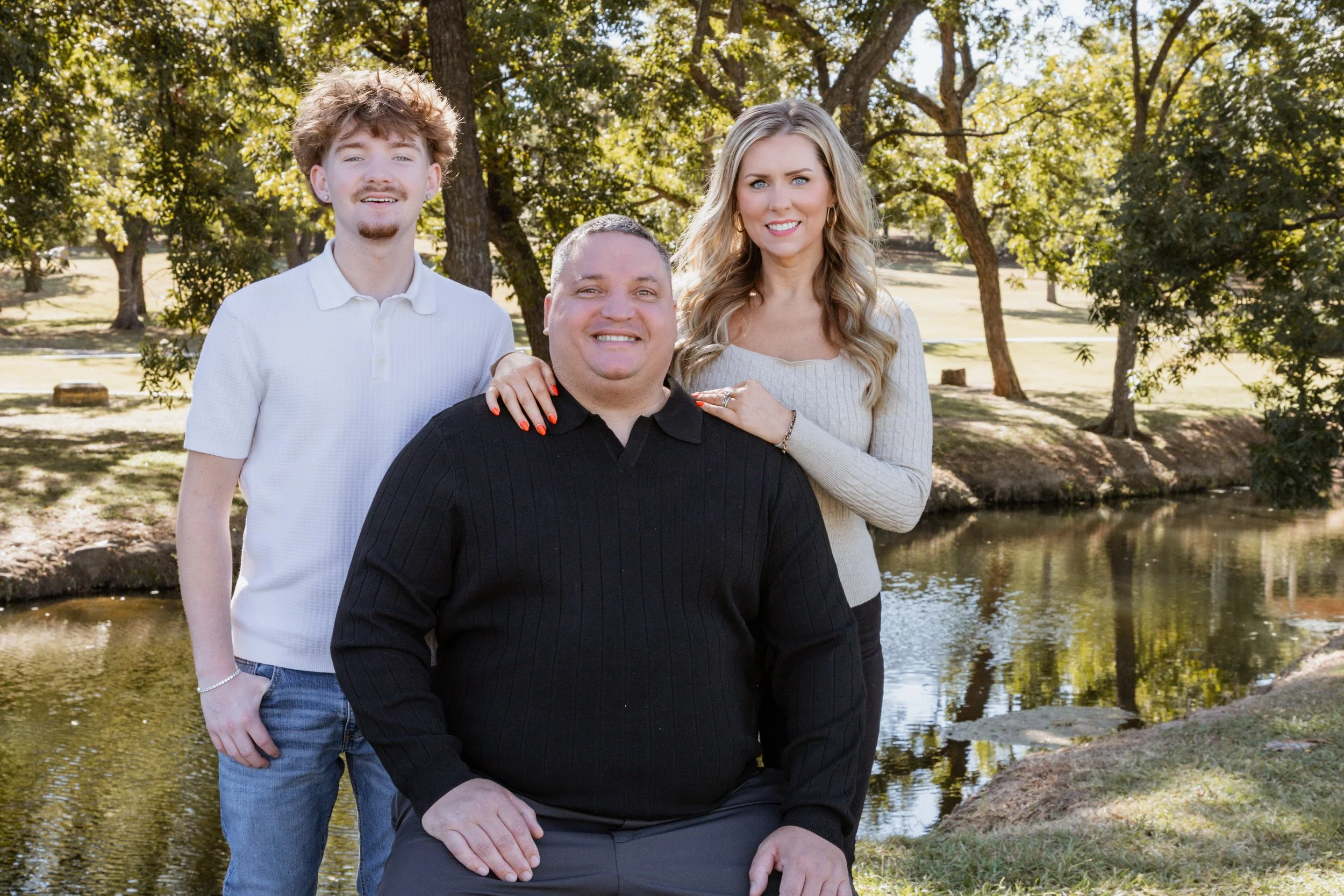 A family of four posing outdoors by a small creek in a park, with trees and grass in the background.