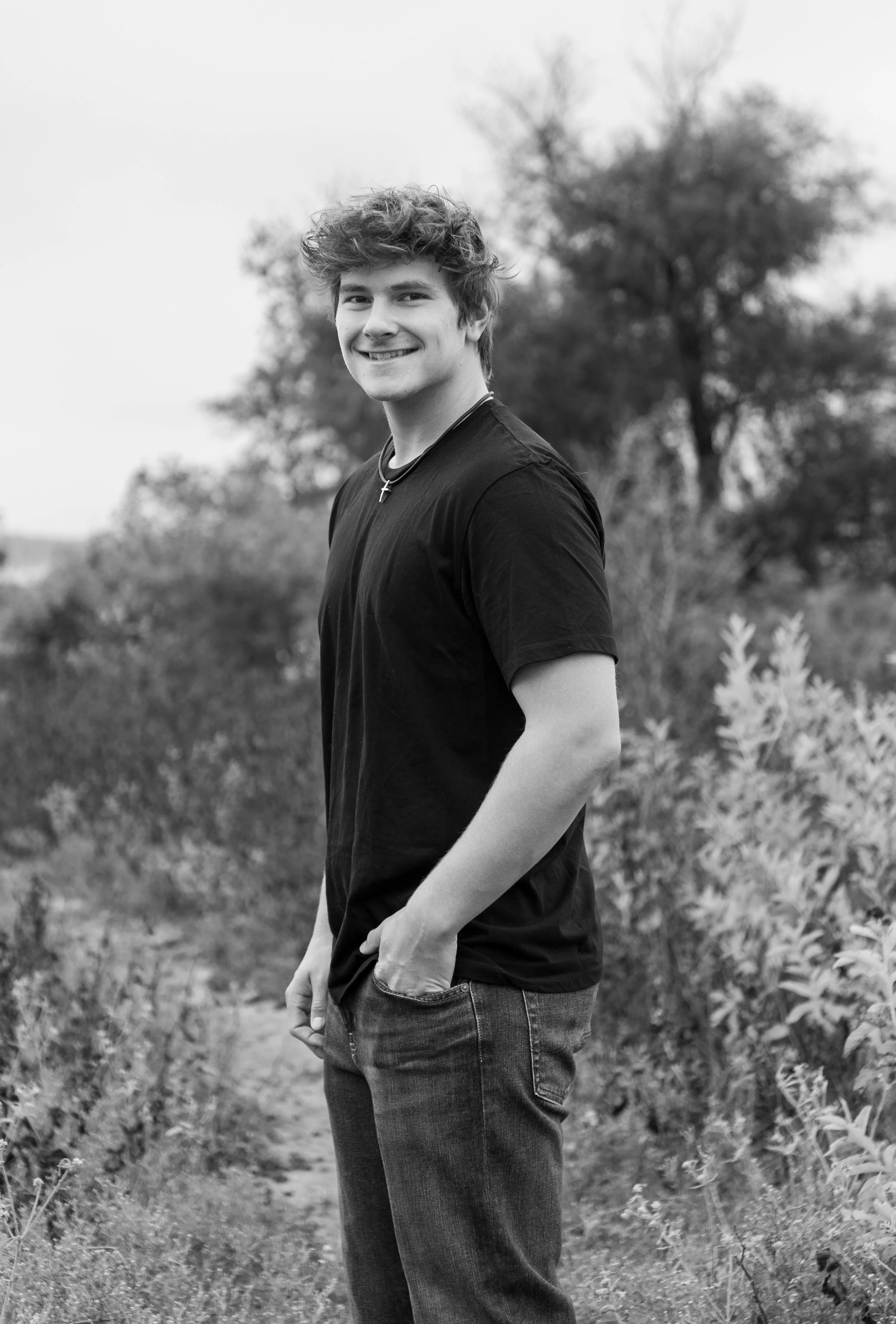 A young man with curly hair smiling and standing outdoors in a natural setting, wearing a black t-shirt and jeans, with trees and bushes in the background.