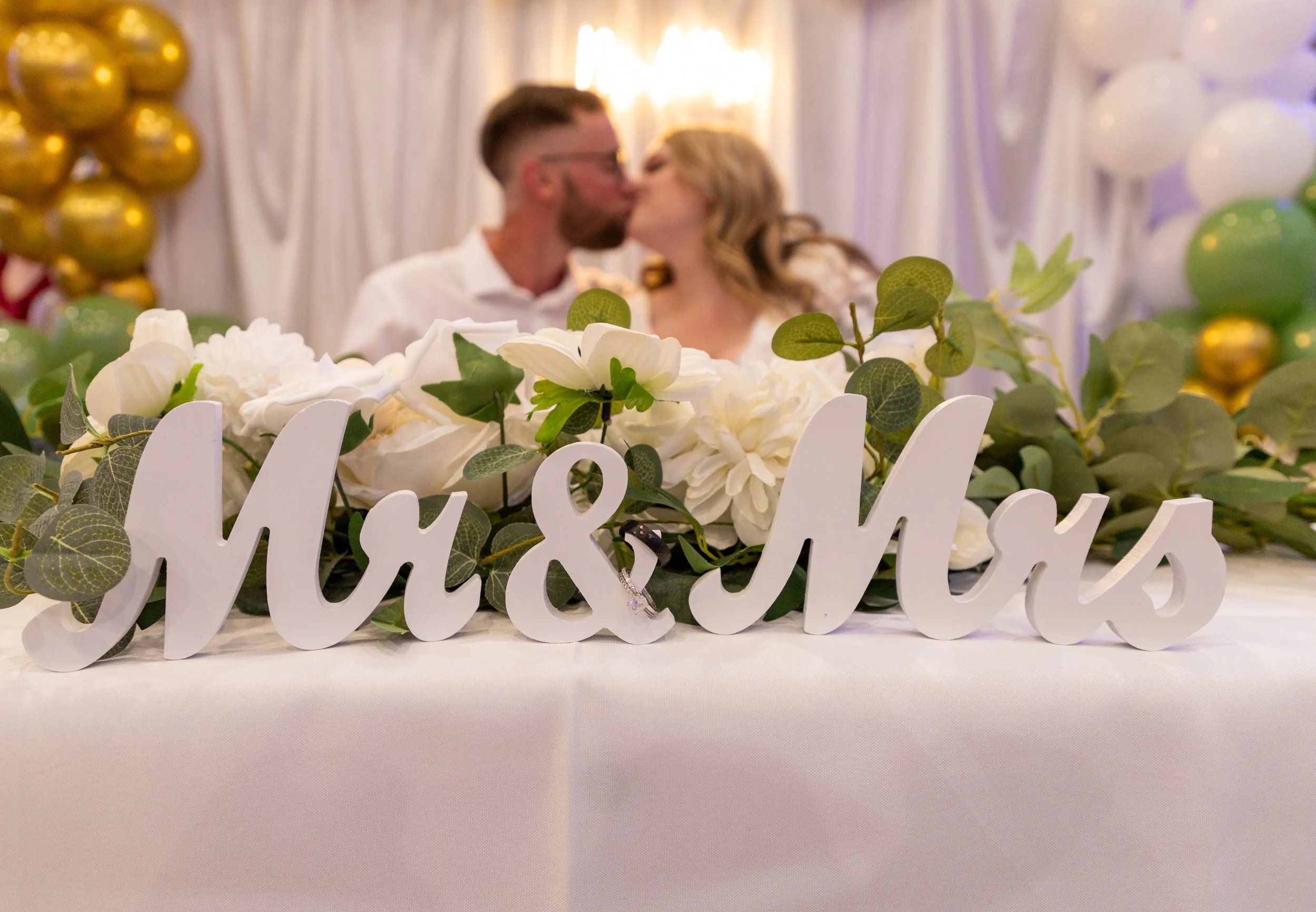 A white table décor with a "Mr & Mrs" sign surrounded by white flowers and greenery, with a blurred background of a couple kissing at a wedding reception.