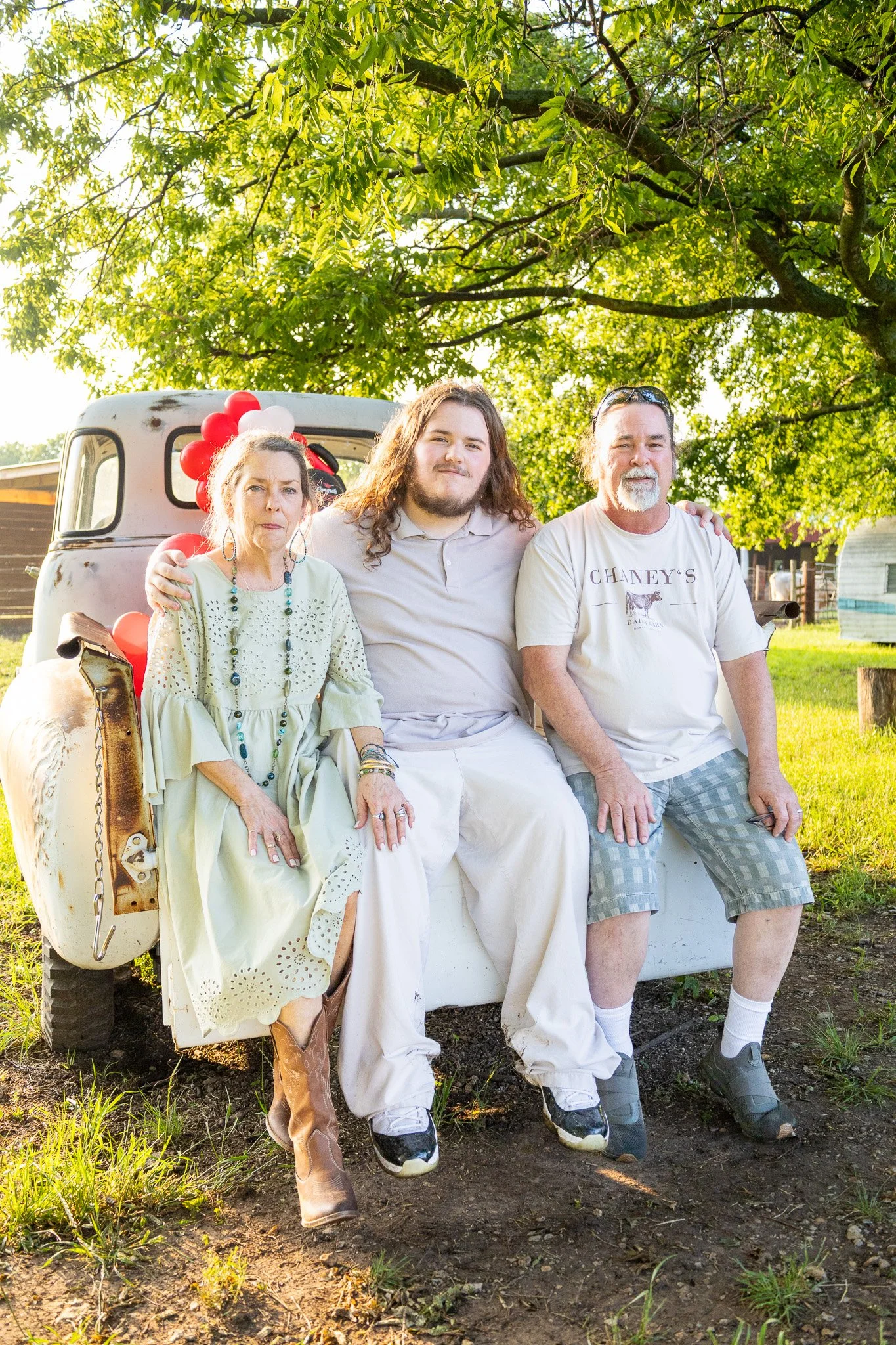 Three people sitting on the back of a vintage truck decorated with red, white, and black balloons under a large leafy tree in a grassy yard.