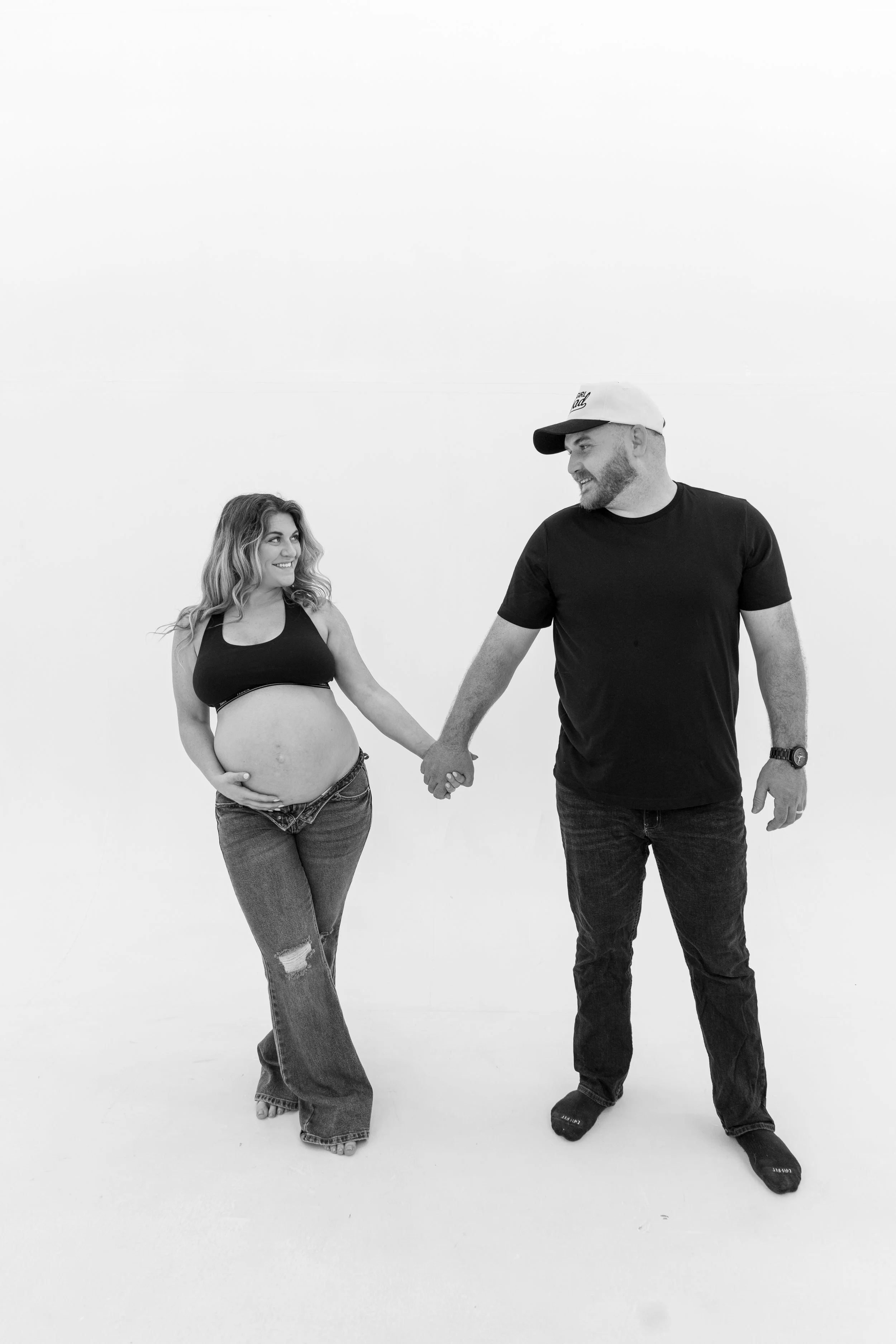 A pregnant woman and a man holding hands and smiling at each other in a minimalistic white studio space.