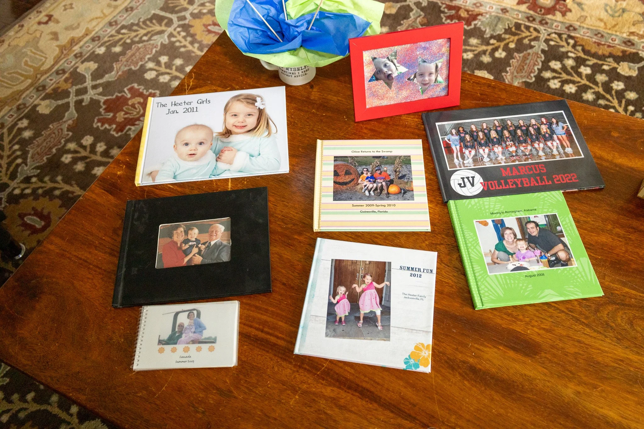 A wooden table holds various photo albums, scrapbooks, and framed pictures, including a Christmas-themed photo, a volleyball team photo, and images of children and family moments.