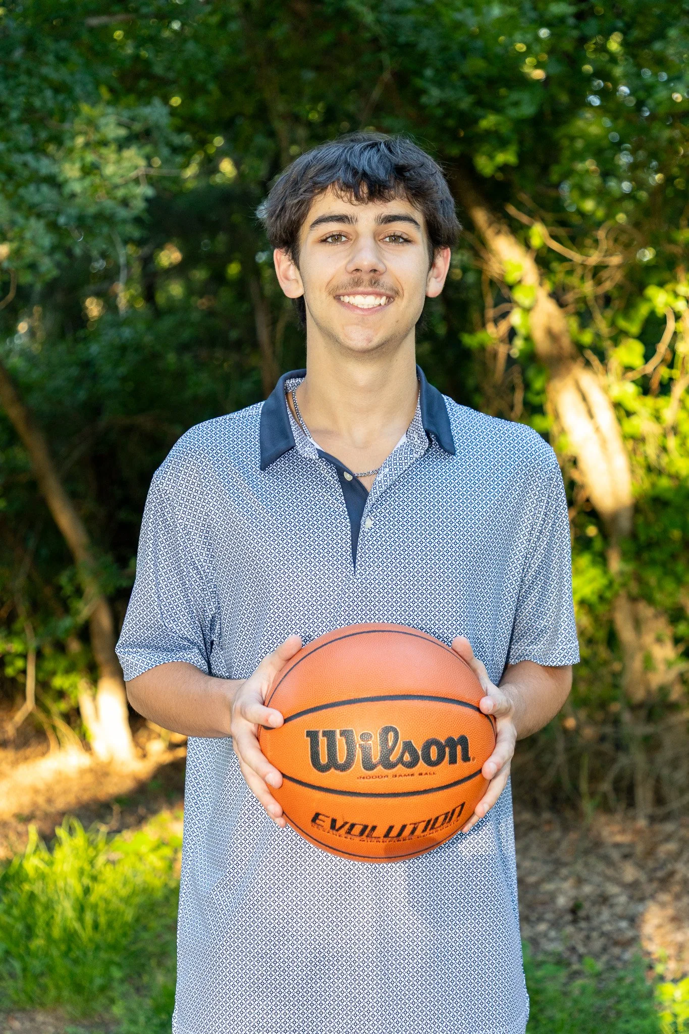 A young man with dark hair, wearing a patterned shirt with a black collar, smiling and holding a Wilson basketball outdoors with green trees in the background.
