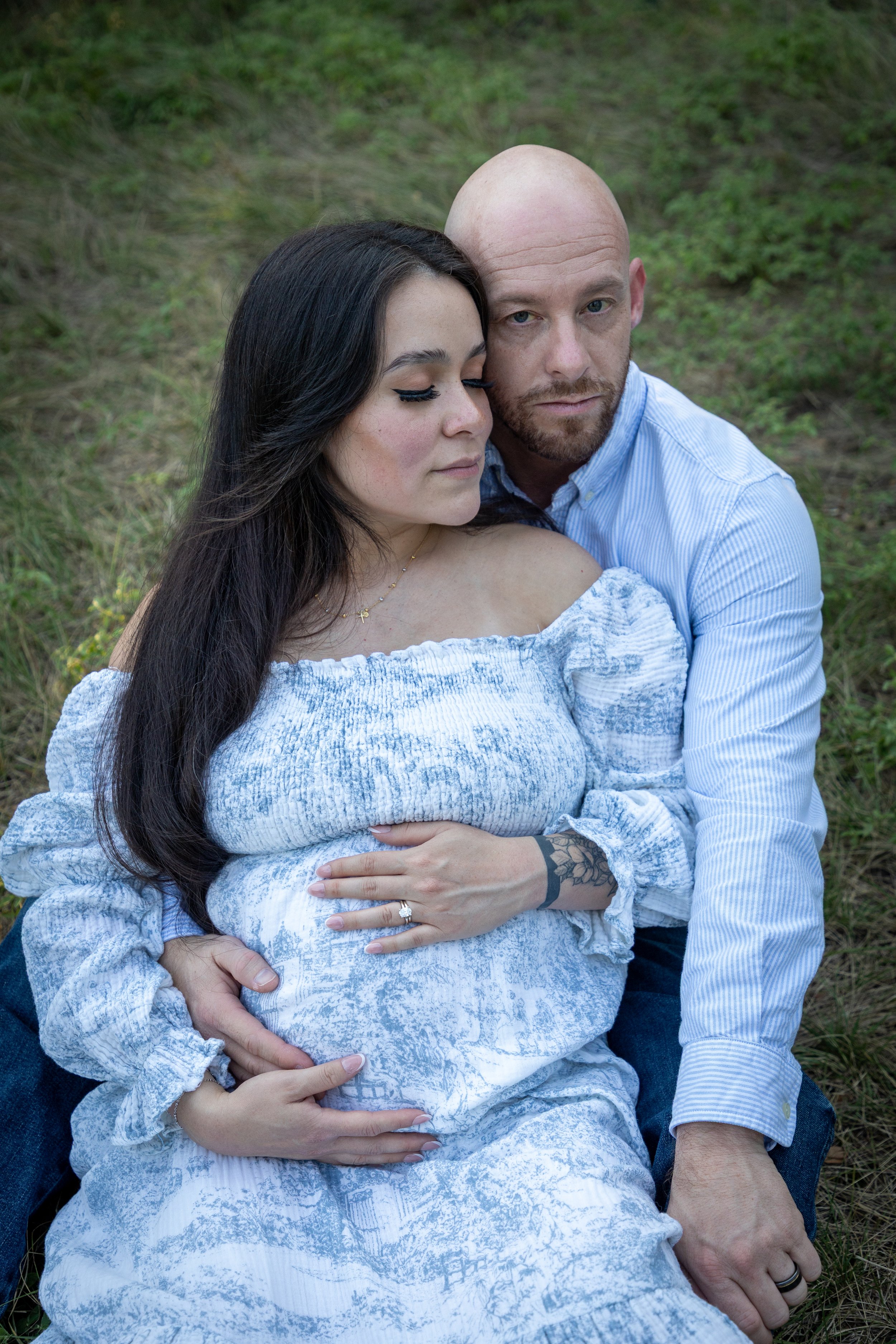 A pregnant woman with long dark hair and a man with a beard and bald head sitting on grass, embracing each other with the woman resting her hand on her belly showing her wedding ring.