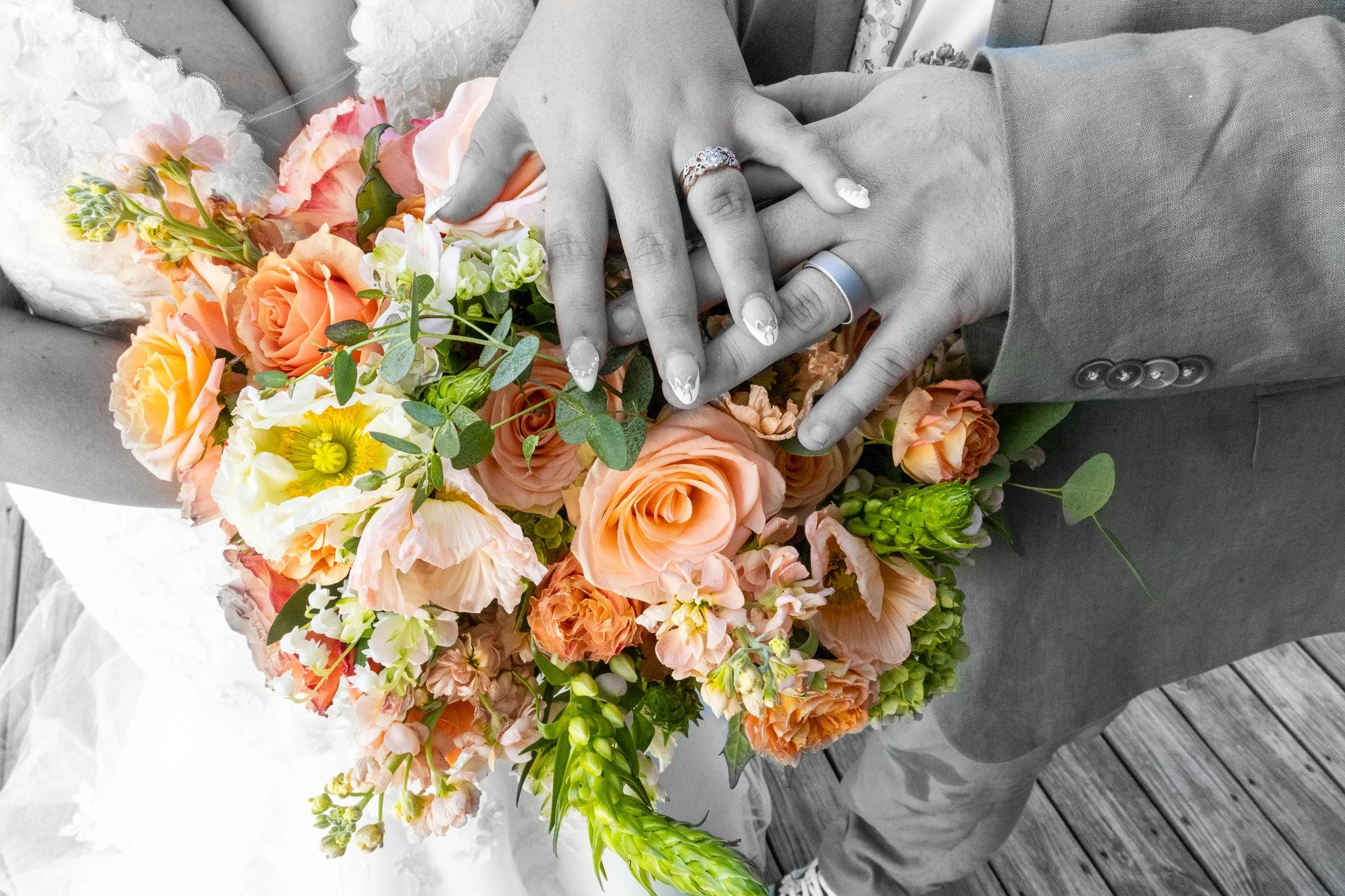 Close-up of a newlywed couple's hands with wedding rings resting on a bouquet of peach, pink, orange, and yellow flowers. The background is in black and white, highlighting the colorful flowers and rings.