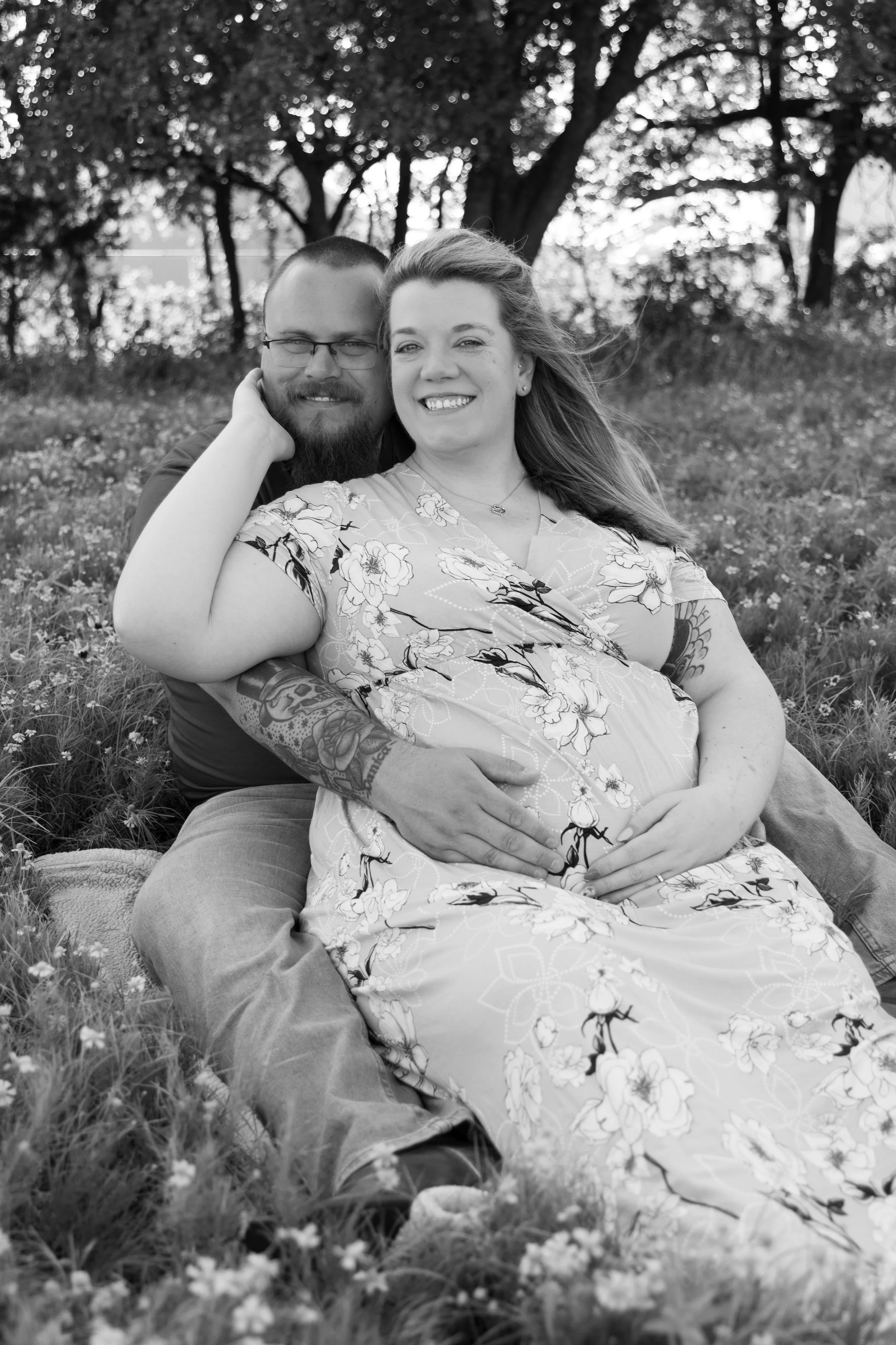 A black and white photo of a smiling couple seated on the grass in a park, with trees in the background. The woman is pregnant, and the man is holding her baby bump.