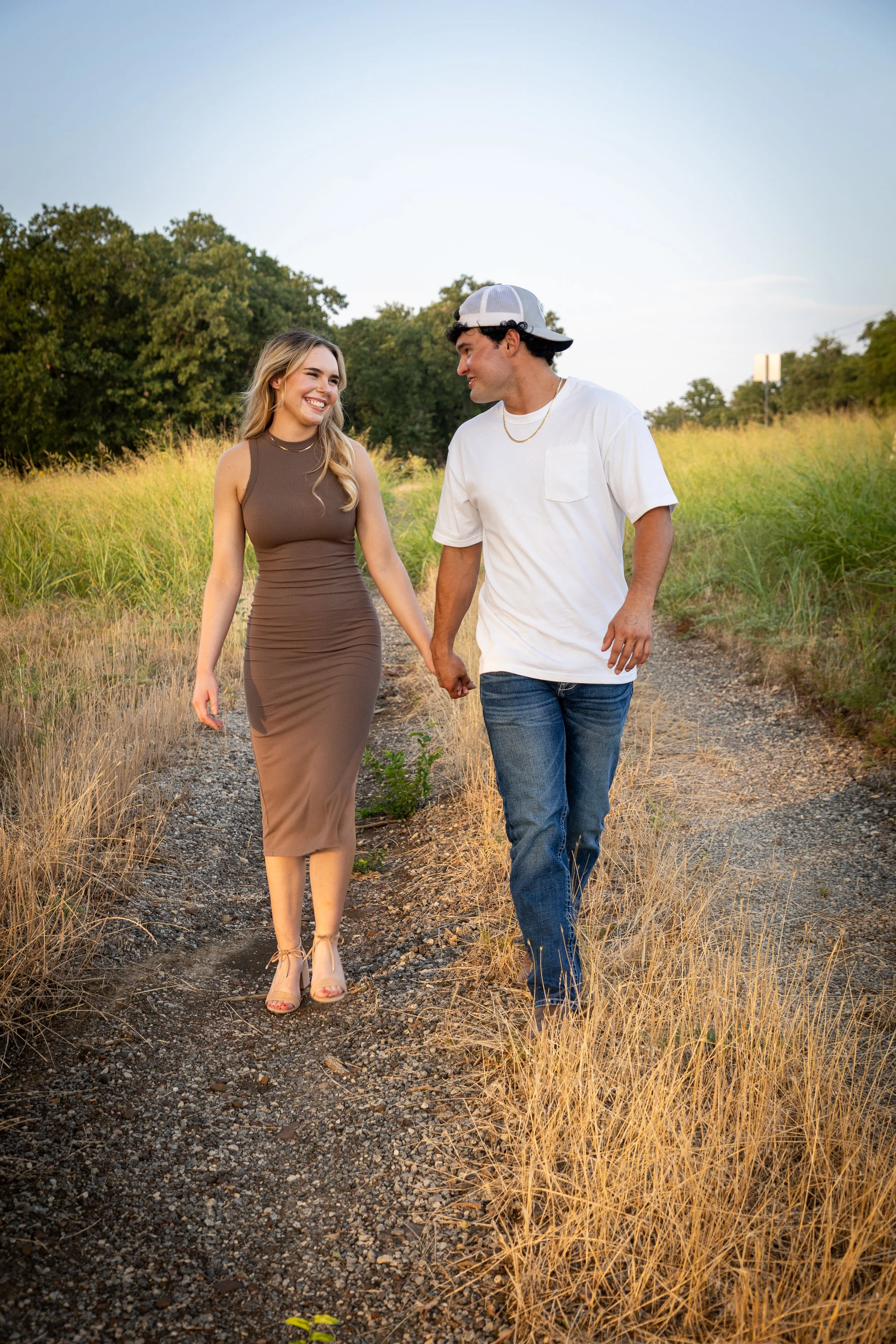 A young man and woman walking hand in hand on a dirt trail through a grassy field, smiling and looking at each other, with trees in the background and clear sky overhead.