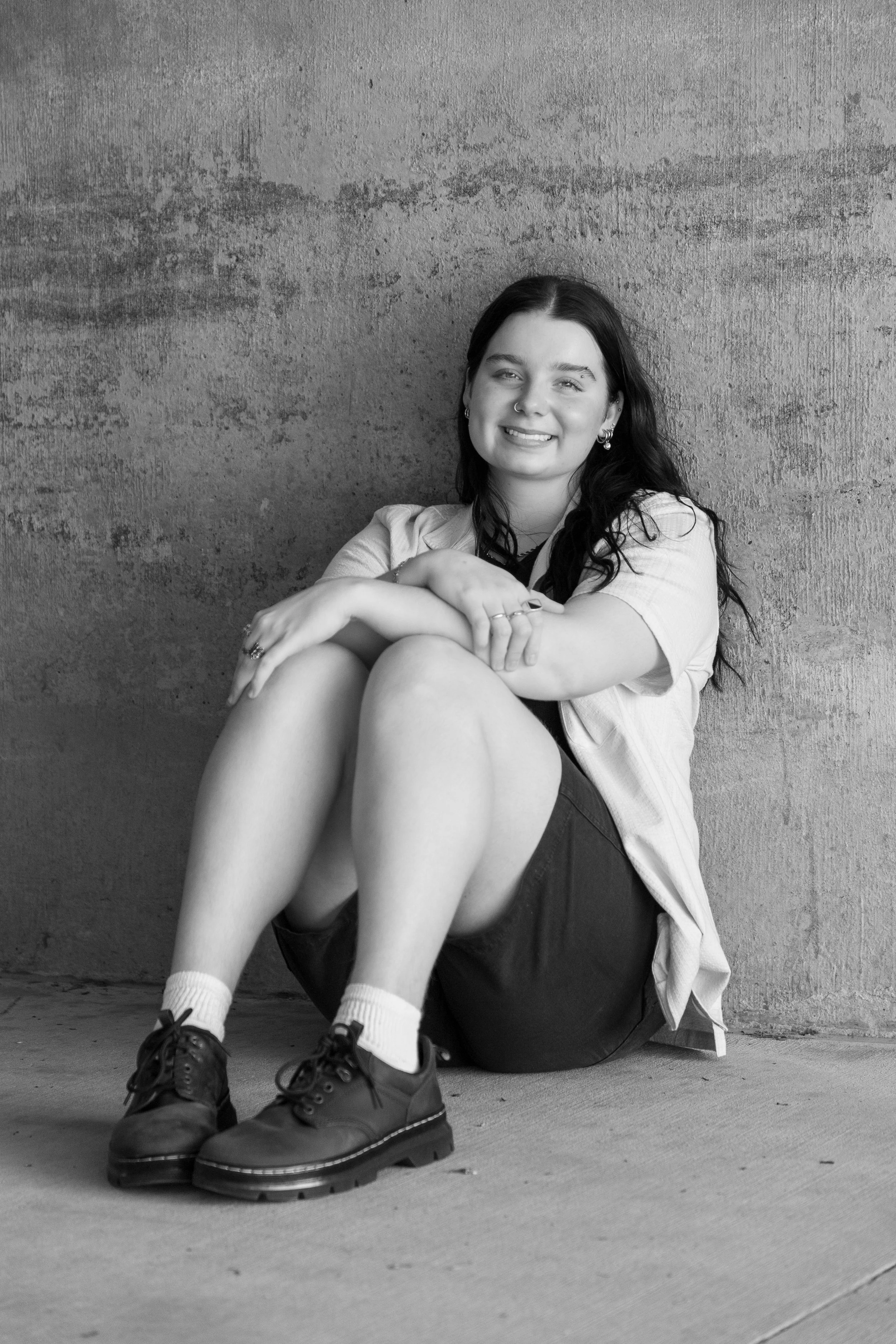 Black and white photo of a young woman sitting against a textured wall, smiling, wearing a casual shirt, shorts, white socks, and boots.