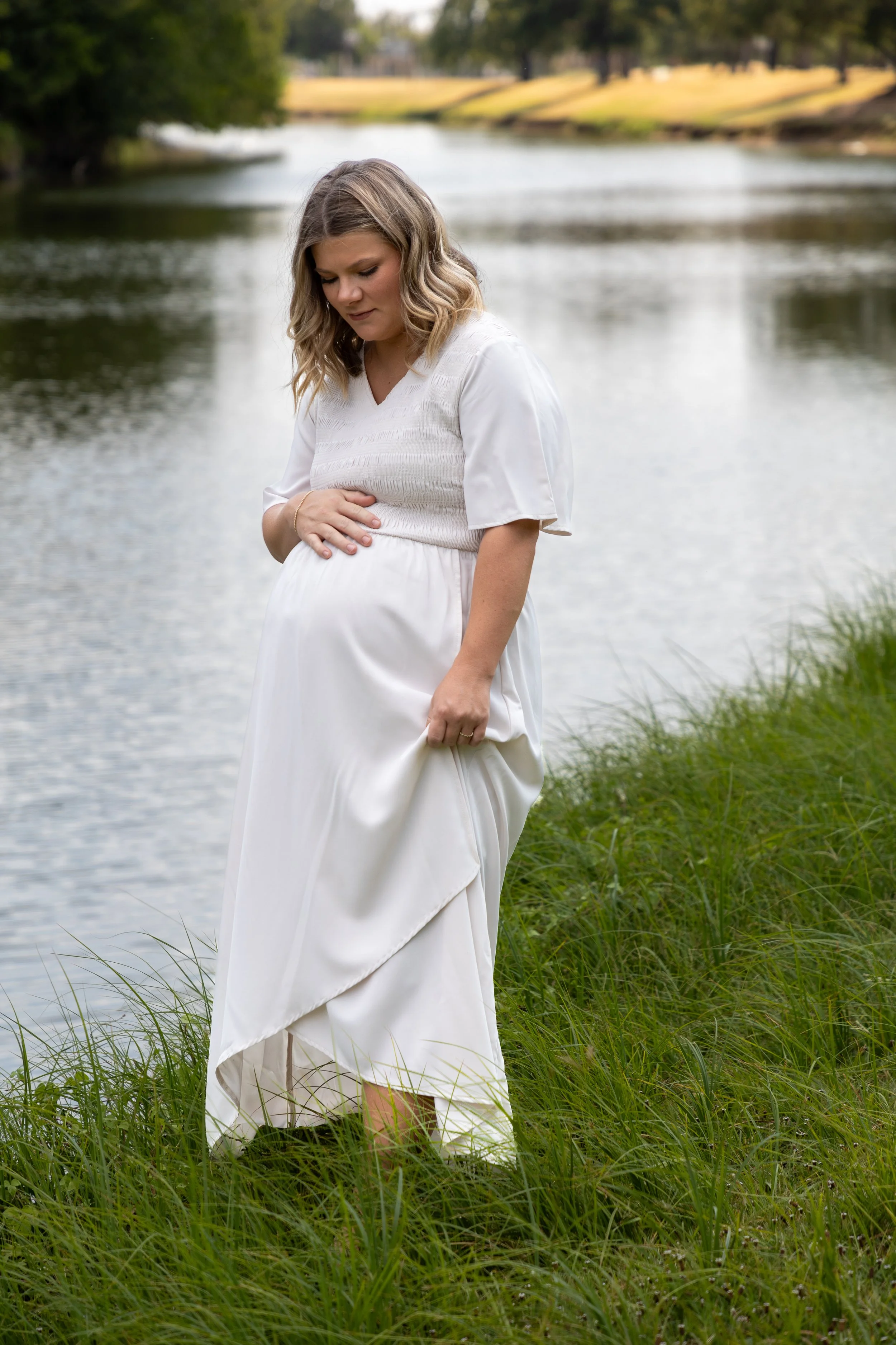 Pregnant woman in a white dress standing on grassy bank near a river, looking down at her belly.