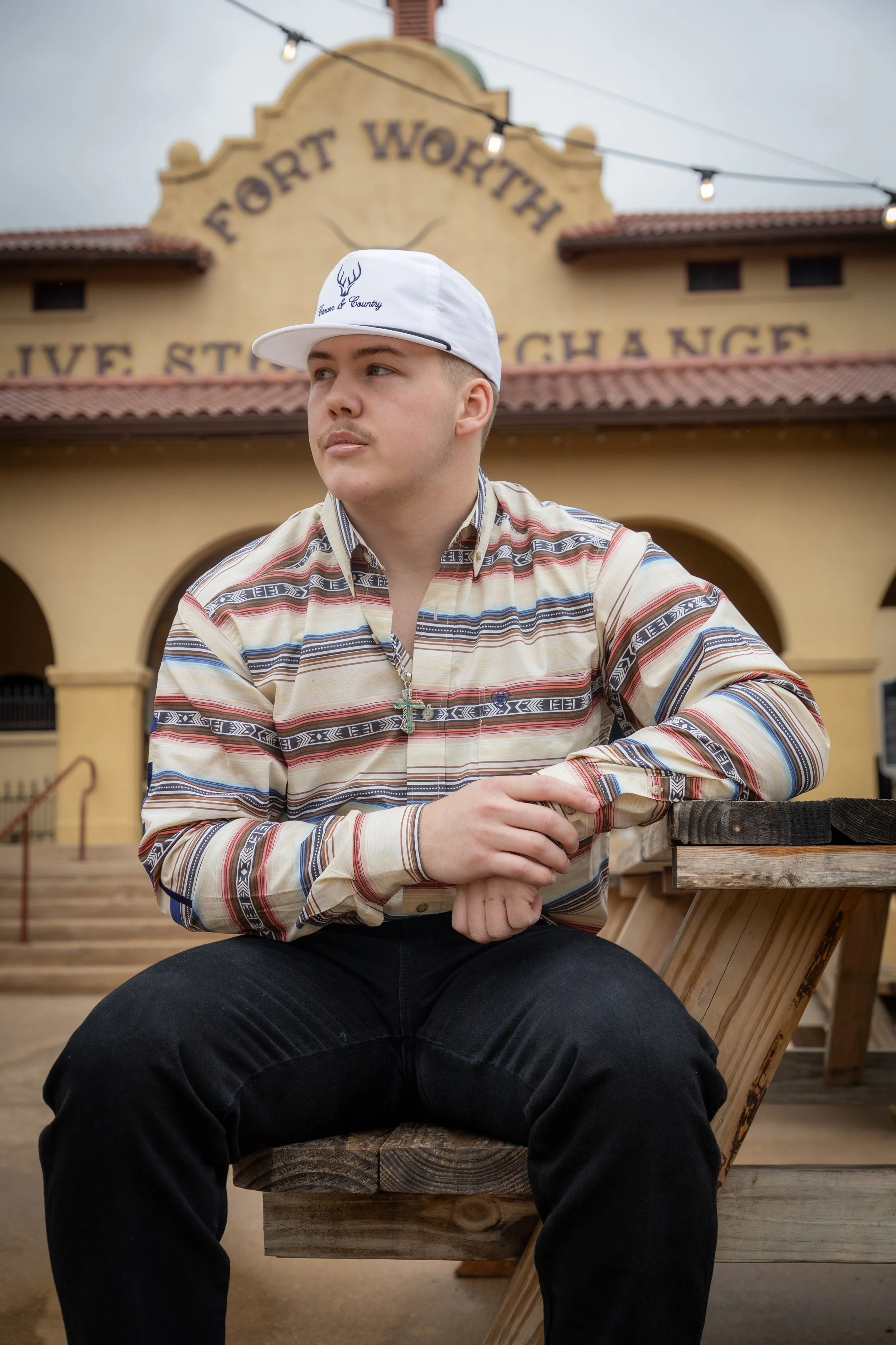 A young man wearing a white cap and a patterned shirt sitting on a wooden bench outside of a yellow building with a sign that reads 'Fort Worth' and 'Live to Change' in the background.