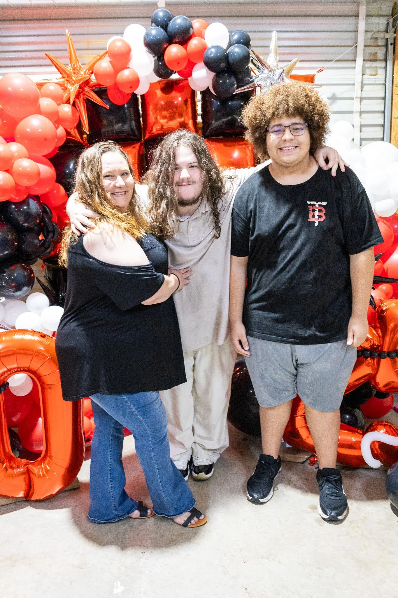 Three people posing in front of a balloon backdrop with red, black, white, and gold balloons and star-shaped balloons, at a celebration or party.