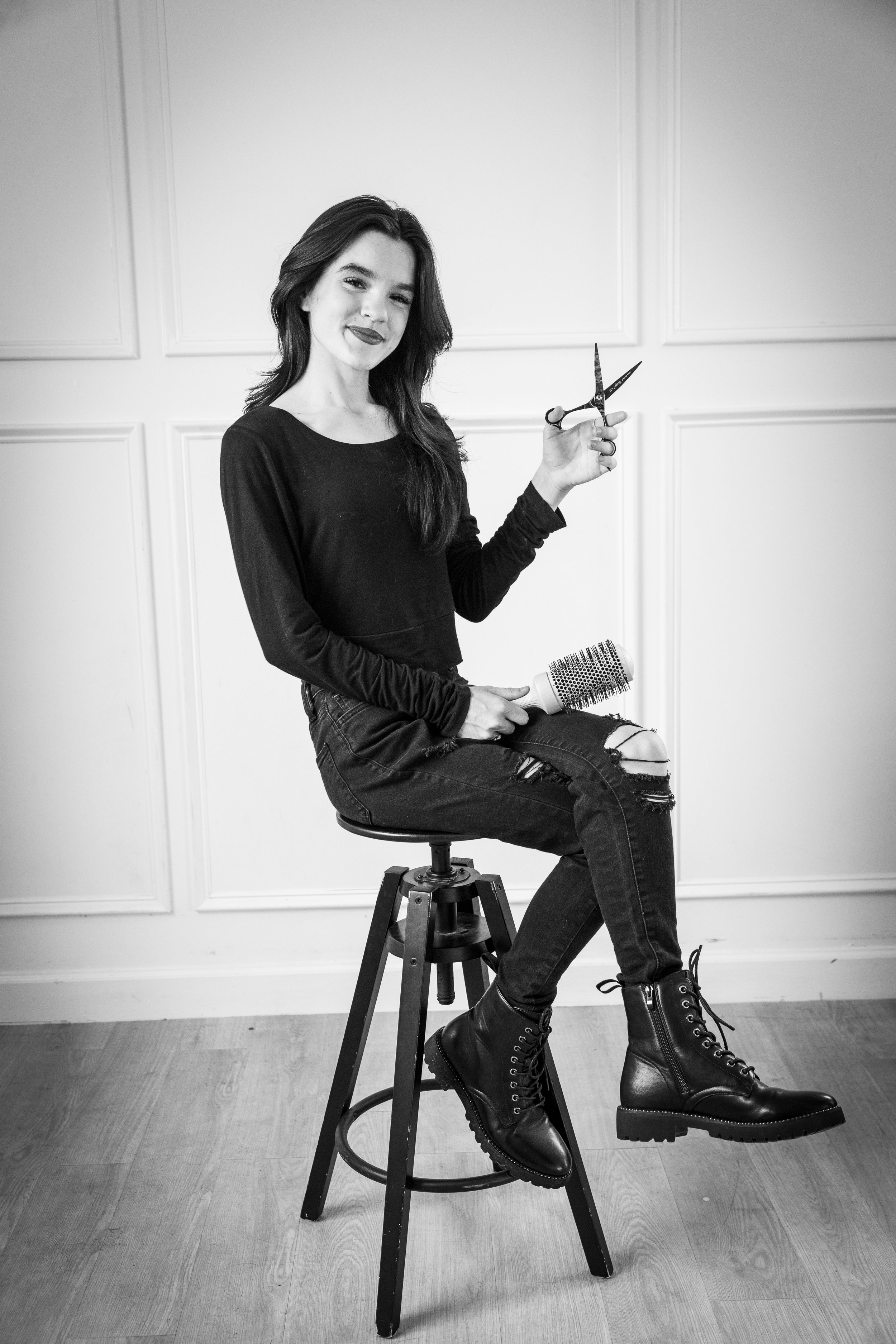 A woman sitting on a stool holding hair scissors and a hairbrush, smiling at the camera, dressed in a black long-sleeve shirt, ripped black jeans, and black boots, in an indoor setting with paneled walls.