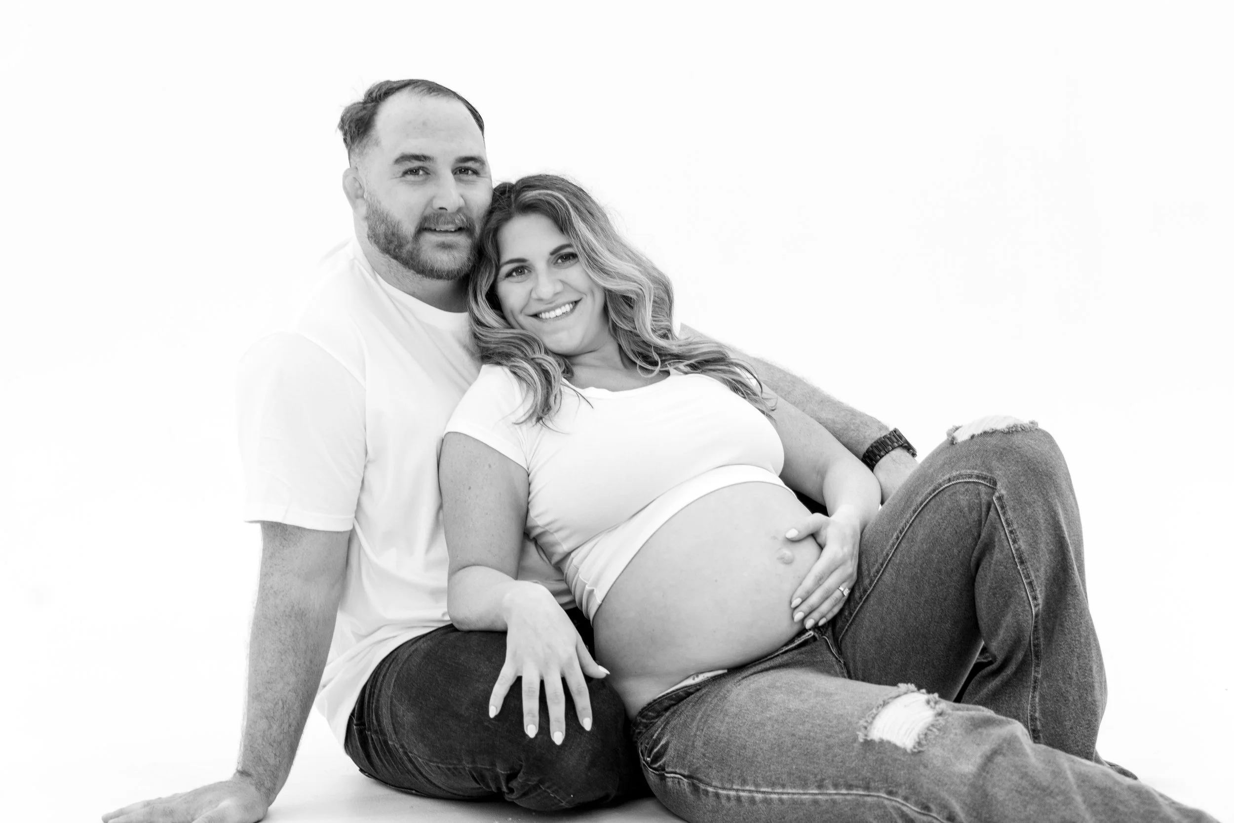 A couple sitting together, with the pregnant woman resting her head on the man's shoulder, both smiling. The woman has long wavy hair and is wearing a white t-shirt and ripped jeans. The man has a beard and short hair.