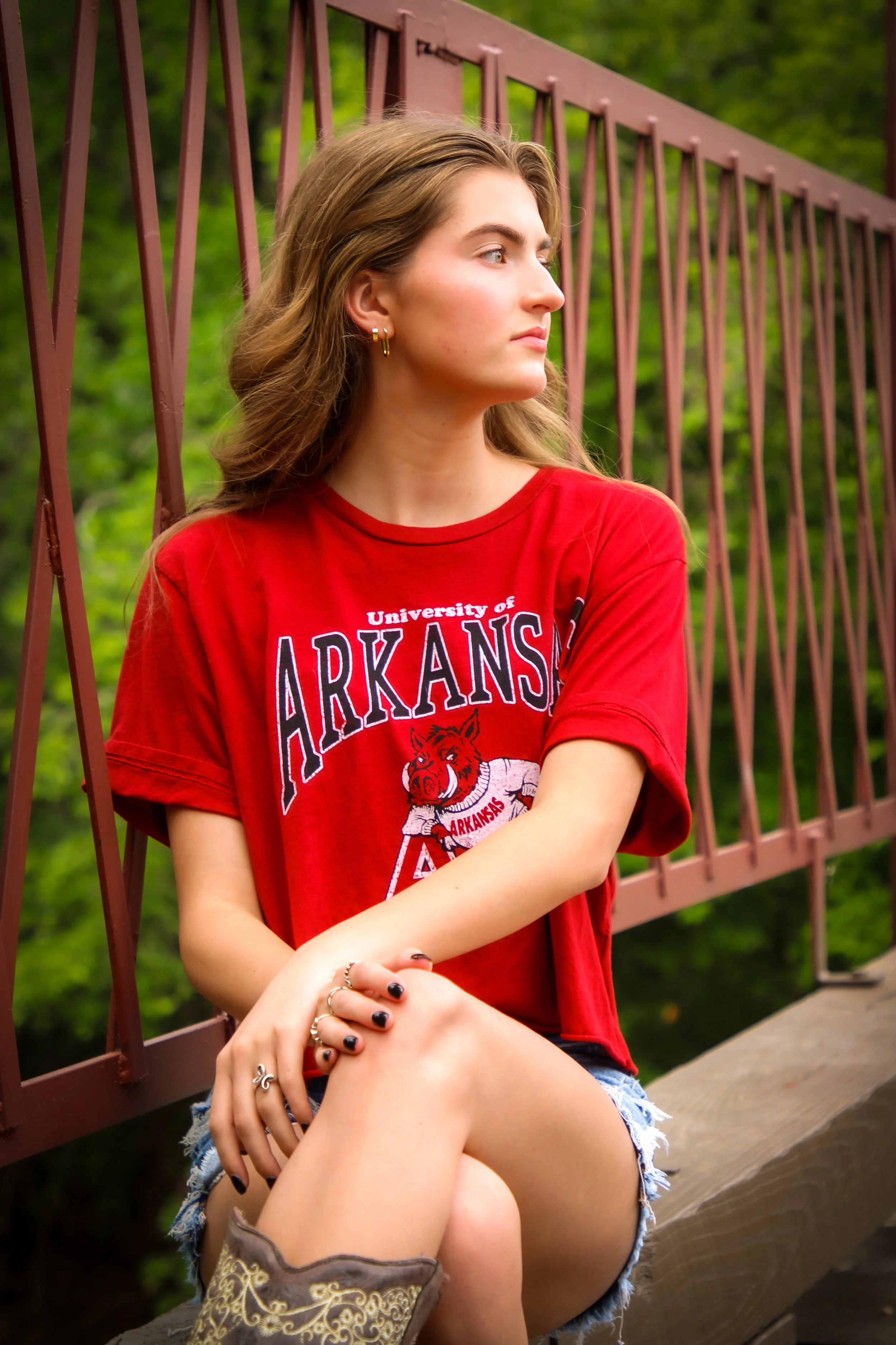 A young woman with long, wavy hair, wearing a red Arkansas T-shirt and denim shorts, sitting on a wooden bench near a metal railing with greenery in the background, looking to the right.