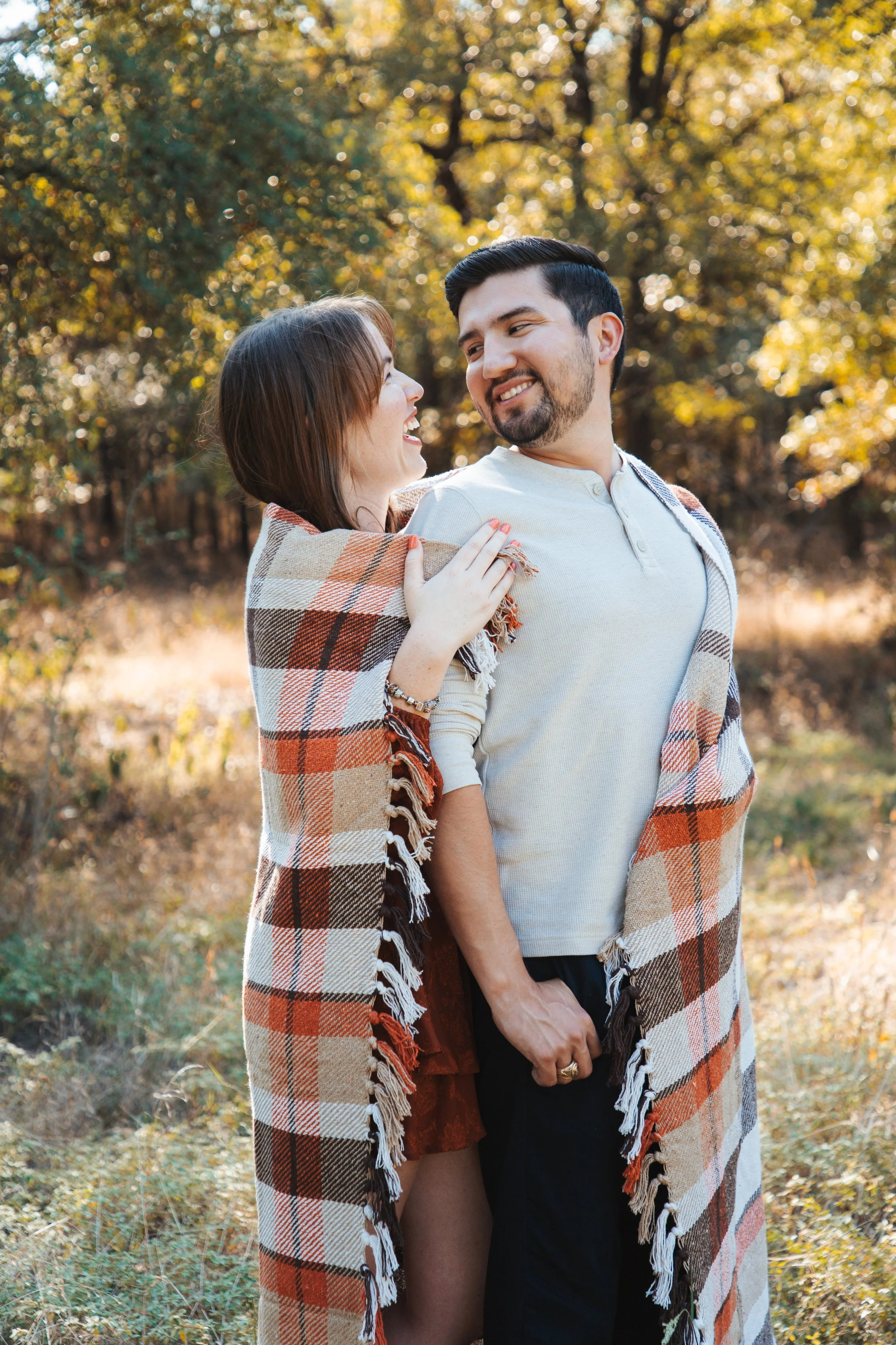 A happy couple standing outdoors in a sunny autumn setting, wrapped in a plaid blanket, smiling at each other.