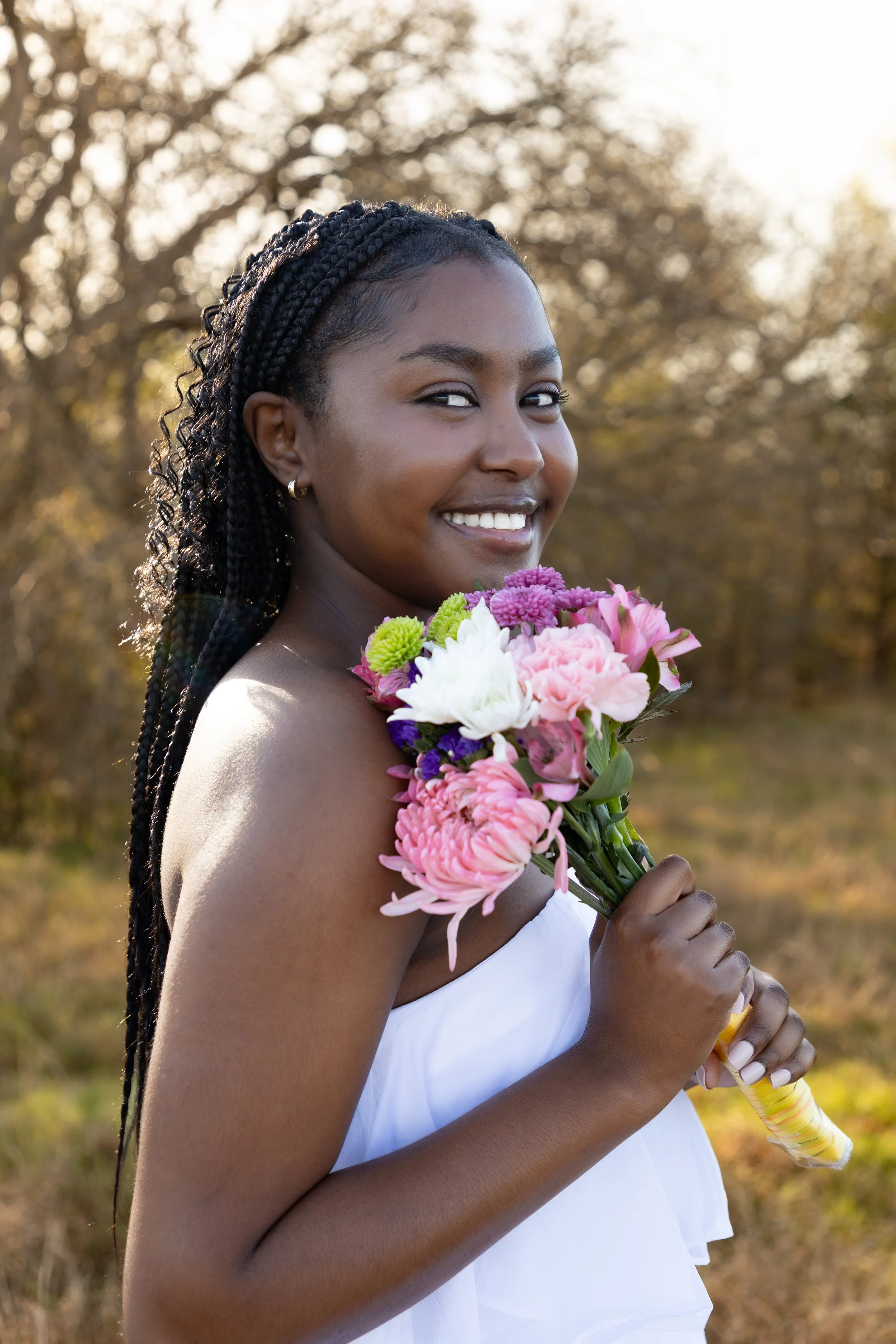 A young woman with braided hair smiling and holding a bouquet of pink, white, and purple flowers outdoors during sunset.