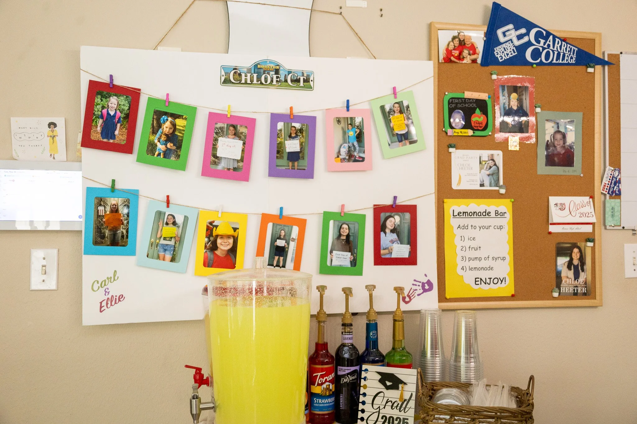 Classroom whiteboard with photographs of children, some holding signs, decorated with colorful paper frames and clips, and college pennants. A lemonade stand with bottles of syrup, cups, and a sign explaining ingredients. A graduation hat and a 2025 
