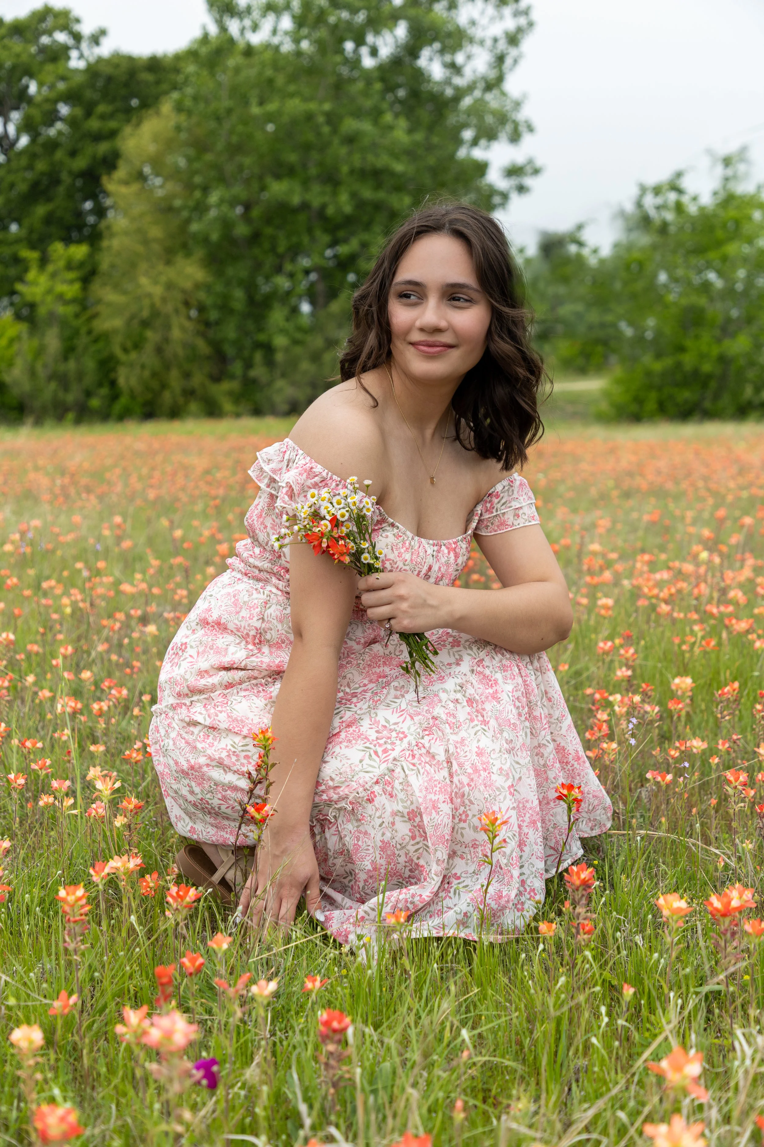 A young woman in a floral pink dress kneels in a flower field, holding a small bouquet of wildflowers, with green trees in the background.