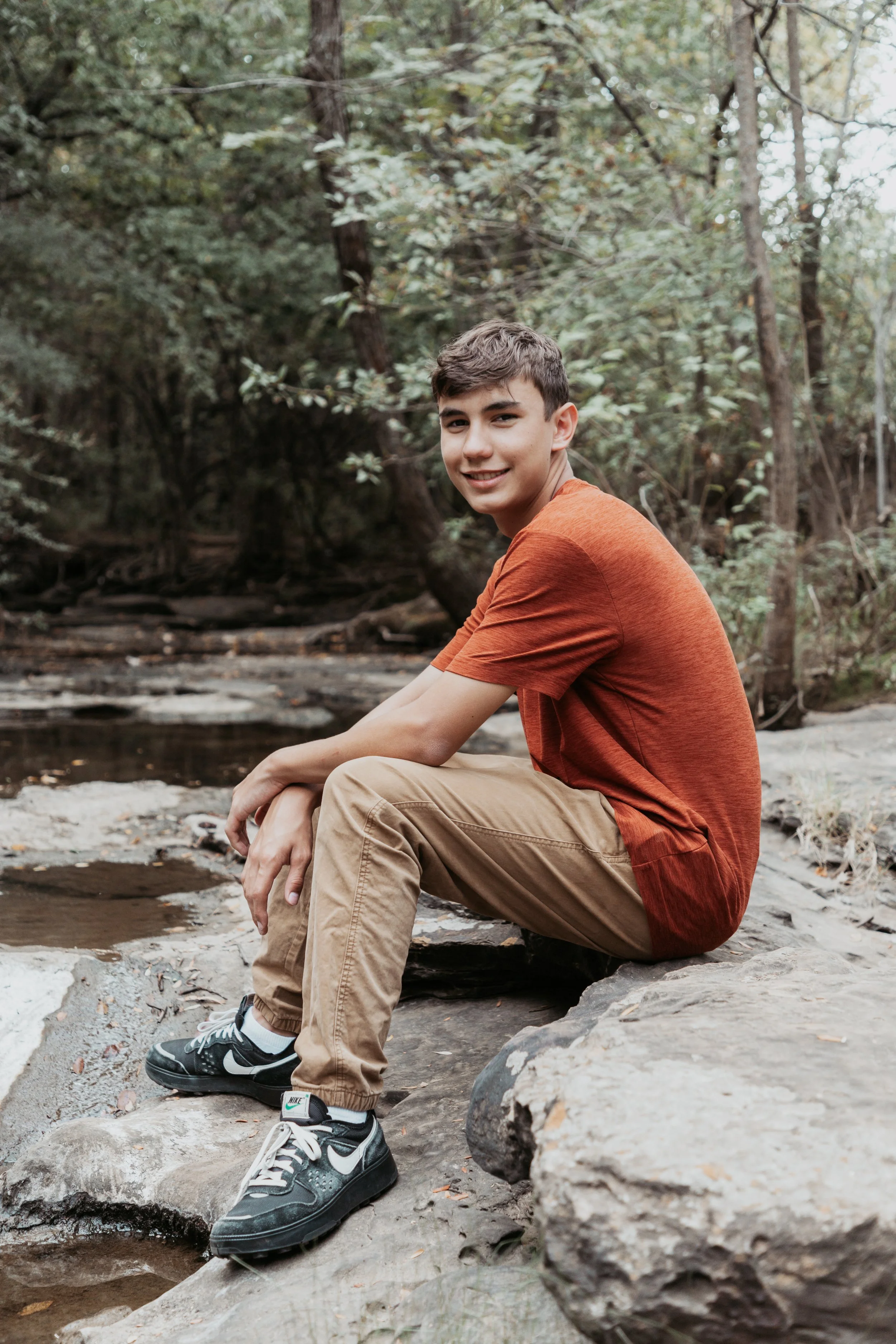 A teenage boy is sitting on rocks next to a creek in a wooded area, smiling at the camera. He is wearing a rust-colored t-shirt, tan pants, and black Nike sneakers.