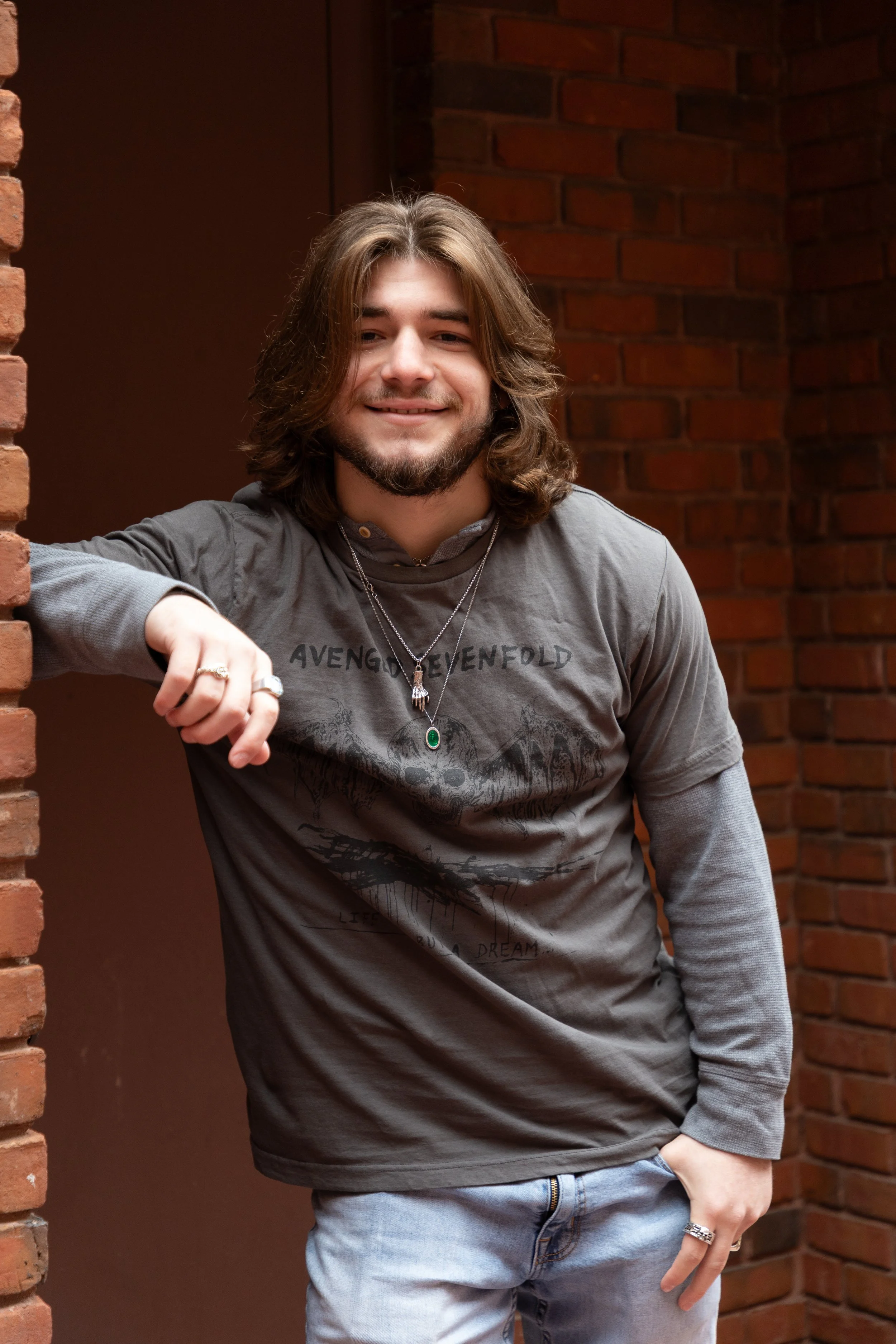 Young man with long brown hair and beard smiling, leaning against a brick wall, wearing a gray T-shirt with black graphics, layered gray long sleeves, and light jeans, in front of a red brick background.