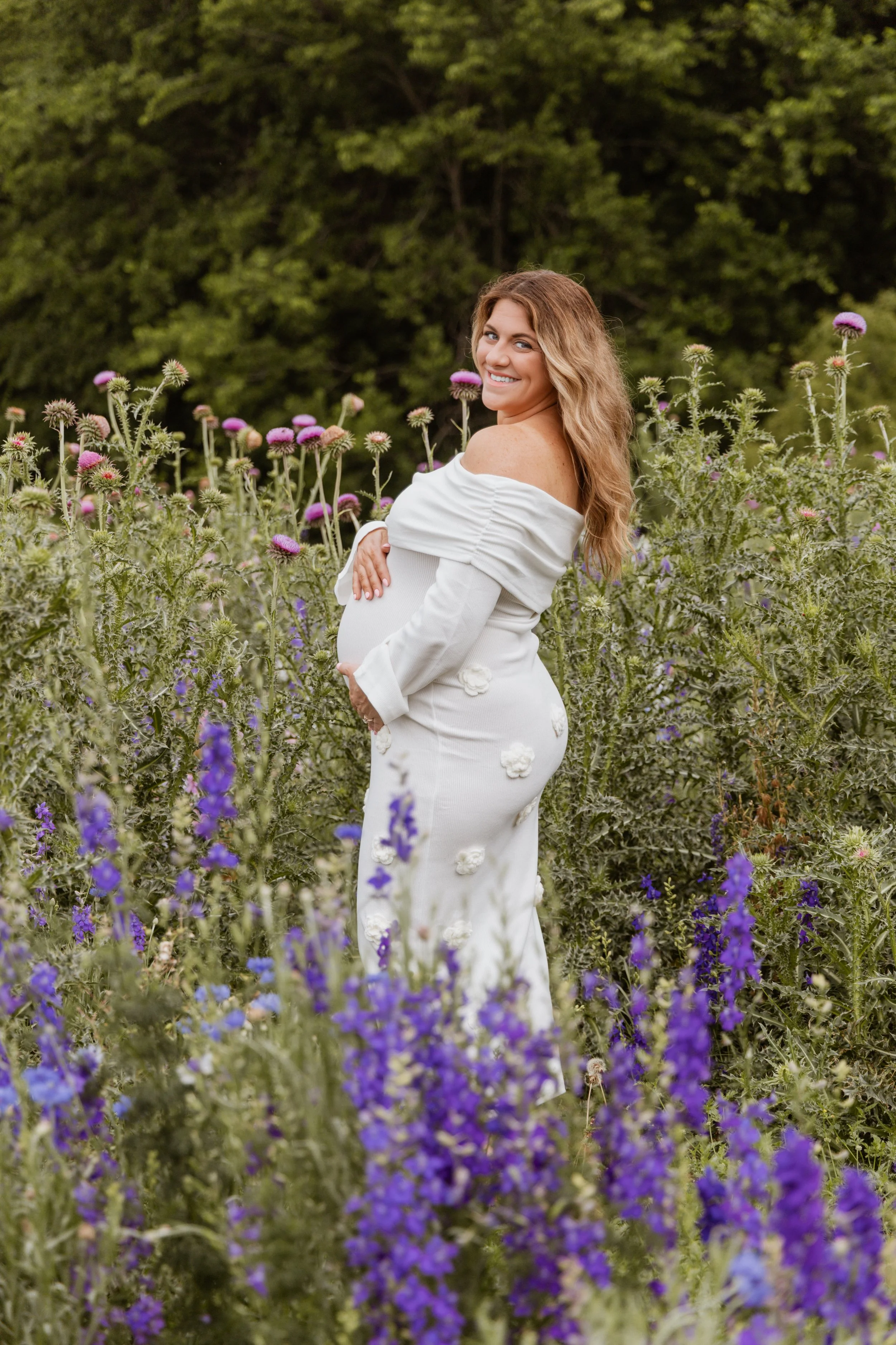 A pregnant woman with long wavy hair smiling and posing among tall purple and pink flowers in a lush green outdoor setting.