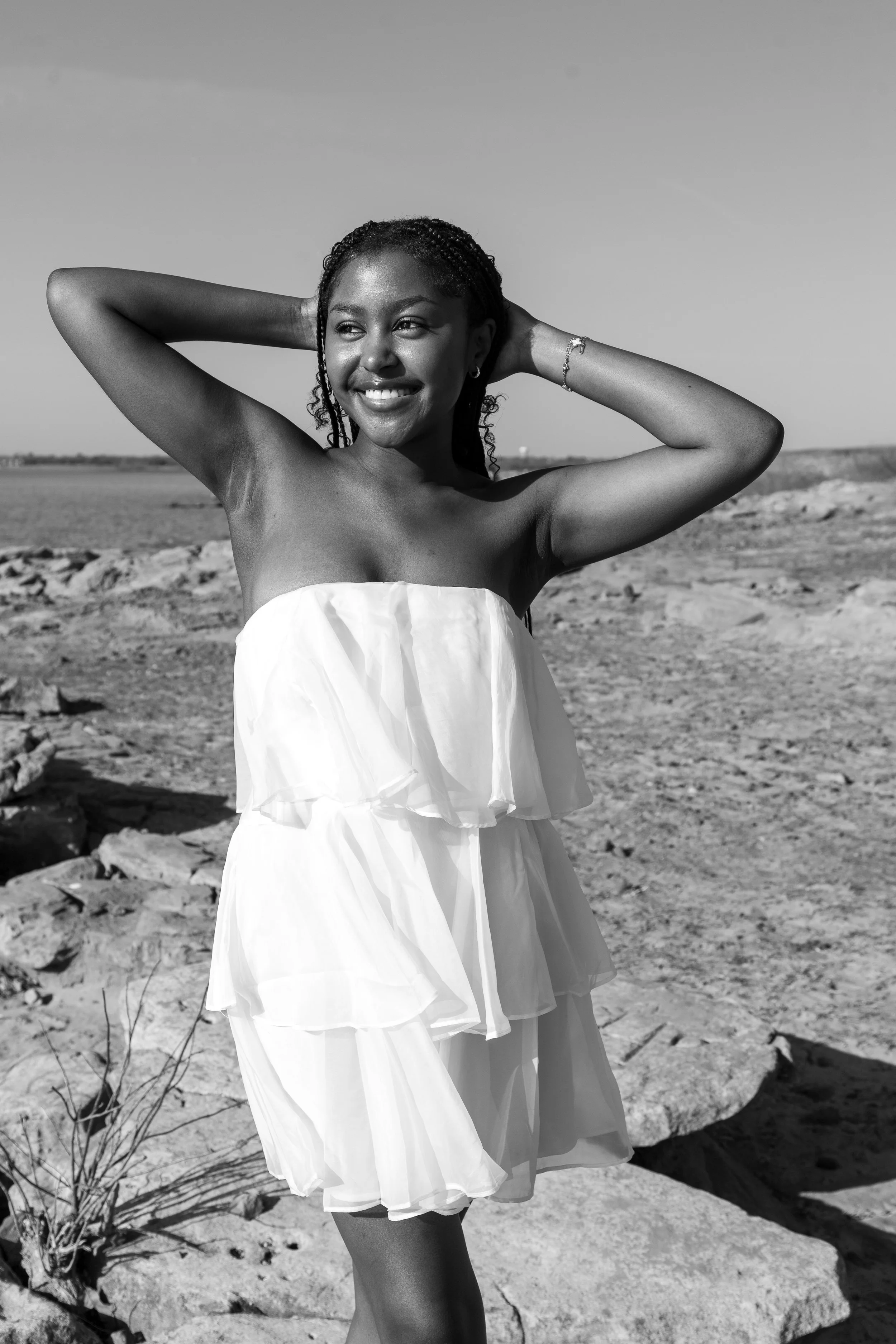A woman smiling with arms raised behind her head in a desert landscape, wearing a strapless white dress.