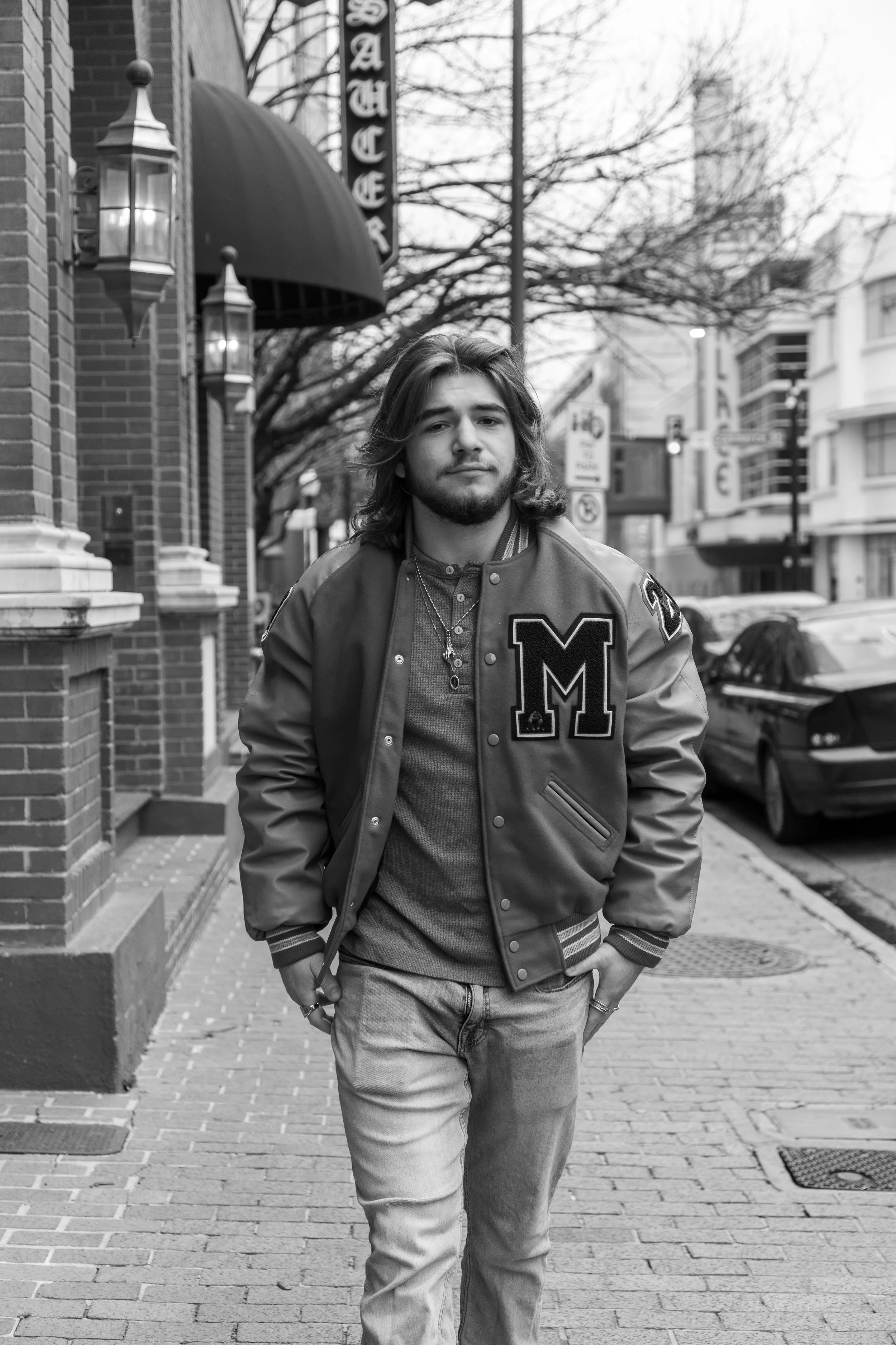 A young man with long hair and a beard walking on a city sidewalk in black and white, wearing a varsity jacket with a large letter 'M' on the chest, jeans, and jewelry, with buildings, parked cars, and street signs in the background.