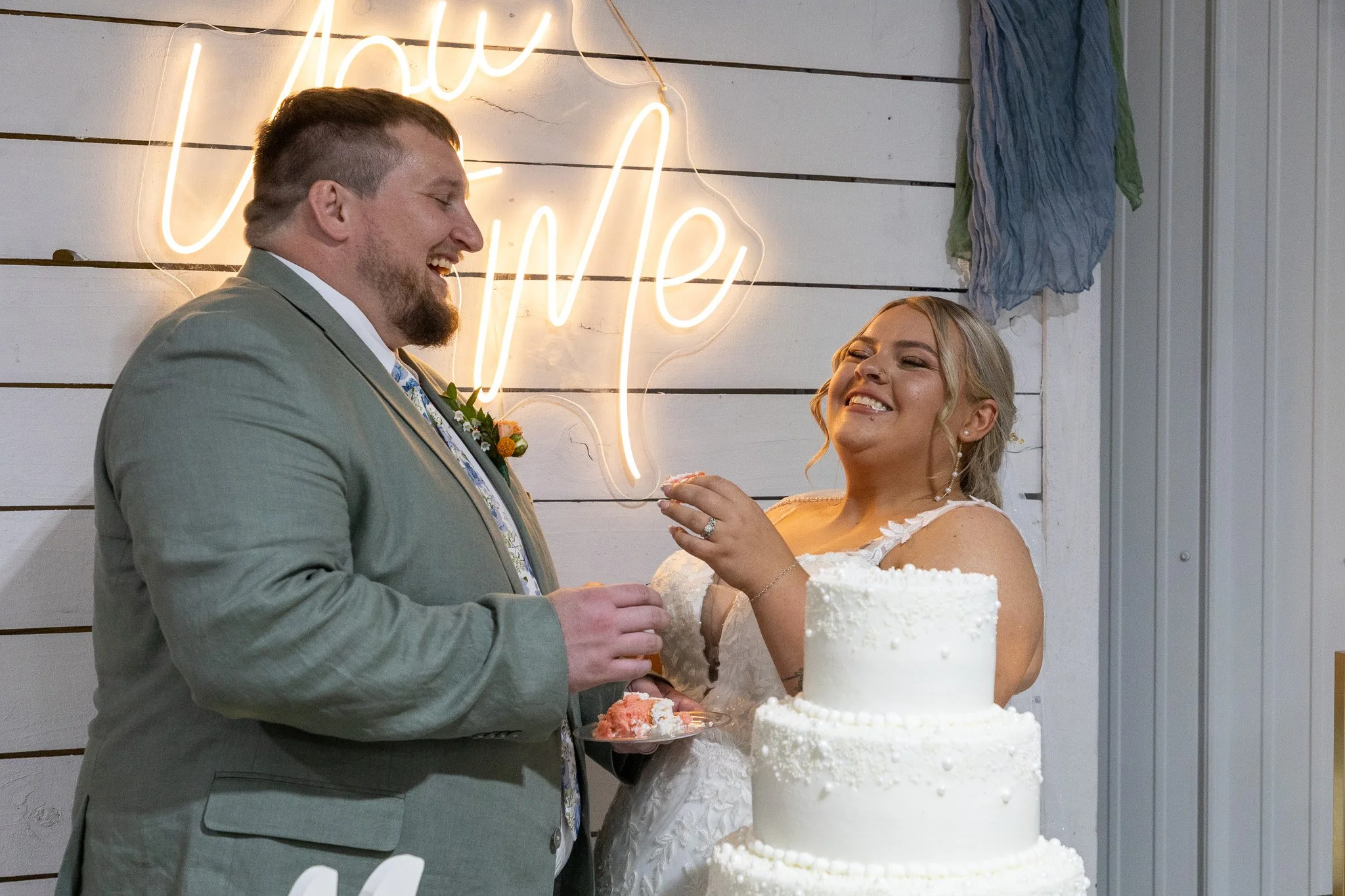 A bride and groom sharing a moment at their wedding reception, standing beside a three-tiered white wedding cake, with a neon sign in the background that says 'love you' on a white wooden wall.